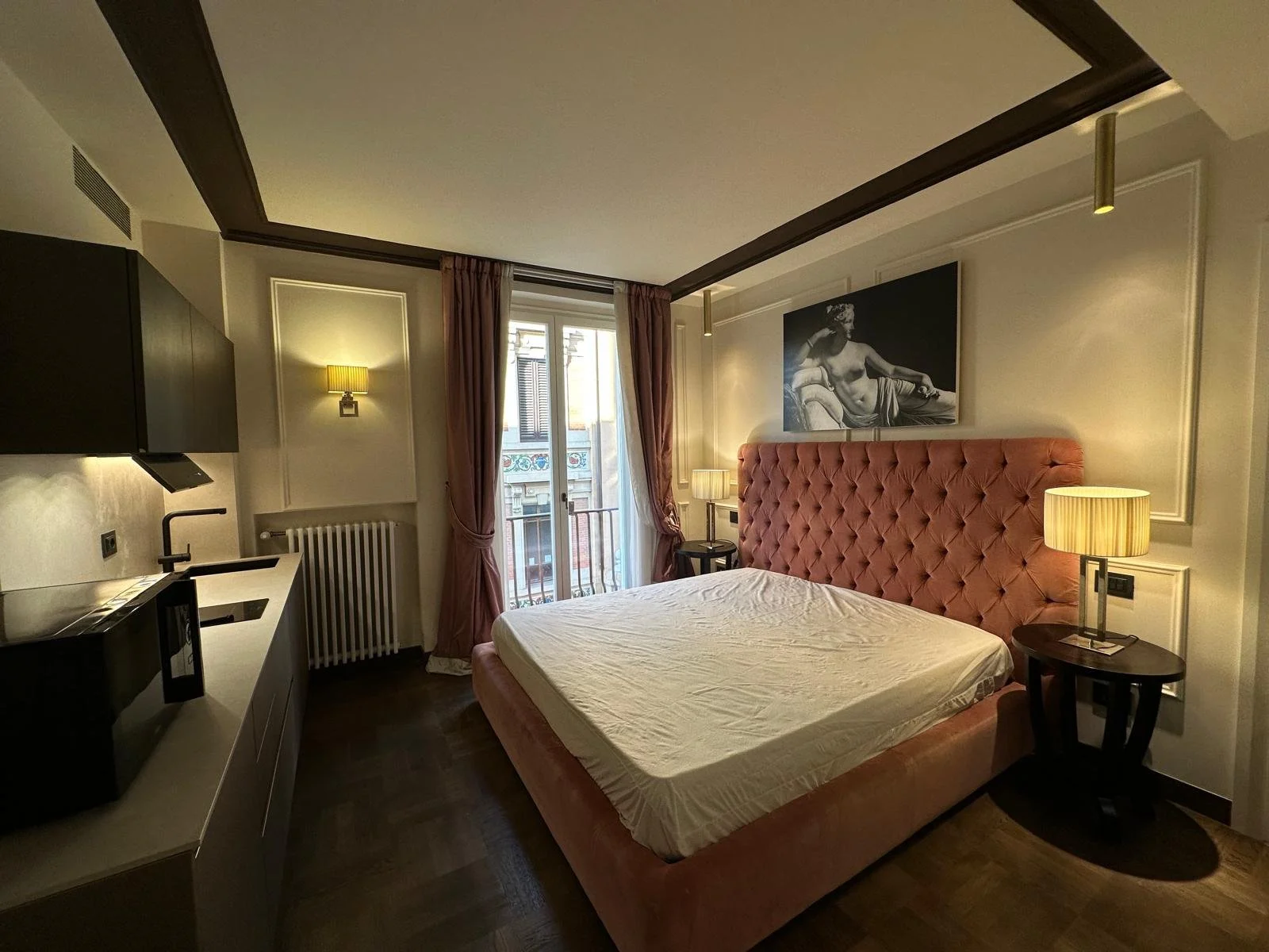 A bedroom with a pink tufted headboard, nightstand, lamps, black and white artwork, a sliding glass door with pink curtains, and a kitchenette area.