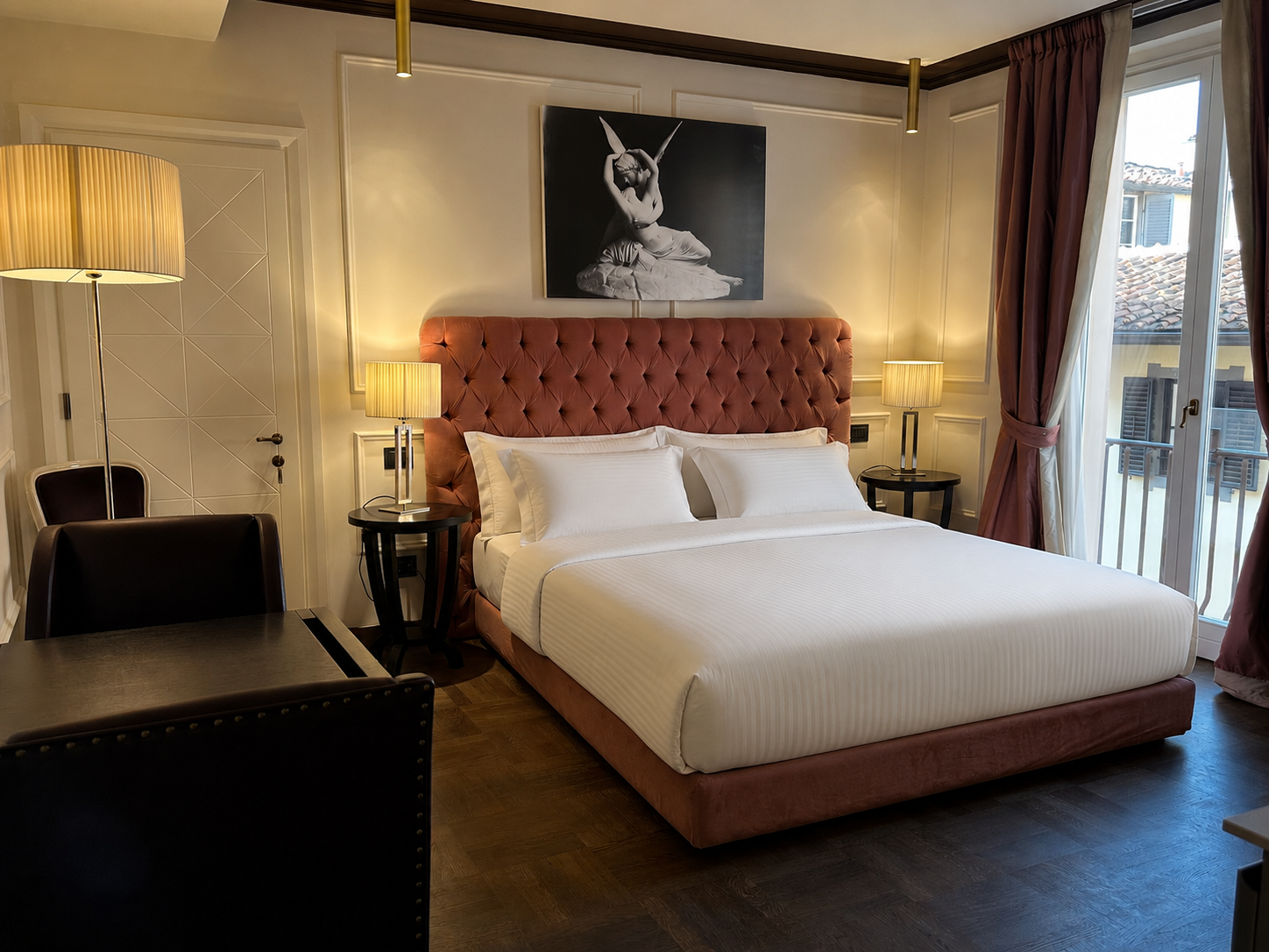A hotel room with a large bed featuring a pink upholstered tufted headboard, flanked by two nightstands with lamps. There is artwork above the bed depicting a ballet dancer. The room has a sliding glass door with pink and white curtains, revealing a 