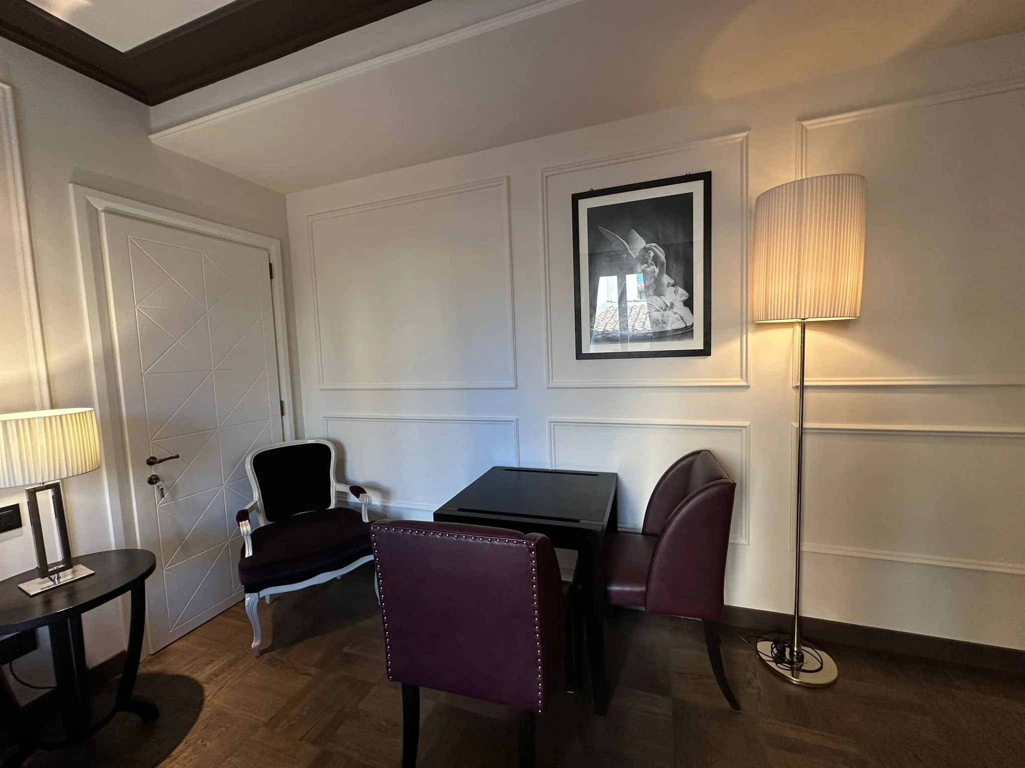 Interior of an elegant room with white paneled walls, black furniture, and a tall floor lamp with a beige shade. There is a framed black-and-white photograph of a woman in a hat on the wall.