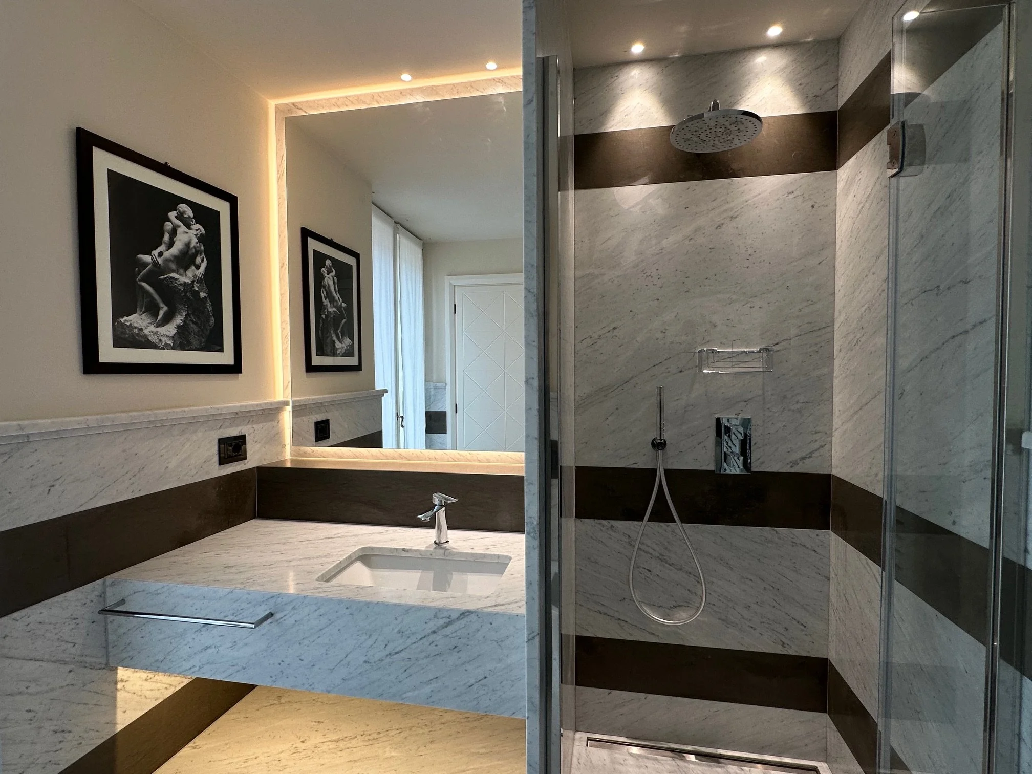 A modern bathroom with marble walls and a shower area with rain showerhead. To the left, there is a window with white curtains and framed black-and-white artwork of a classical sculpture. Recessed ceiling lights illuminate the space, and there is a m