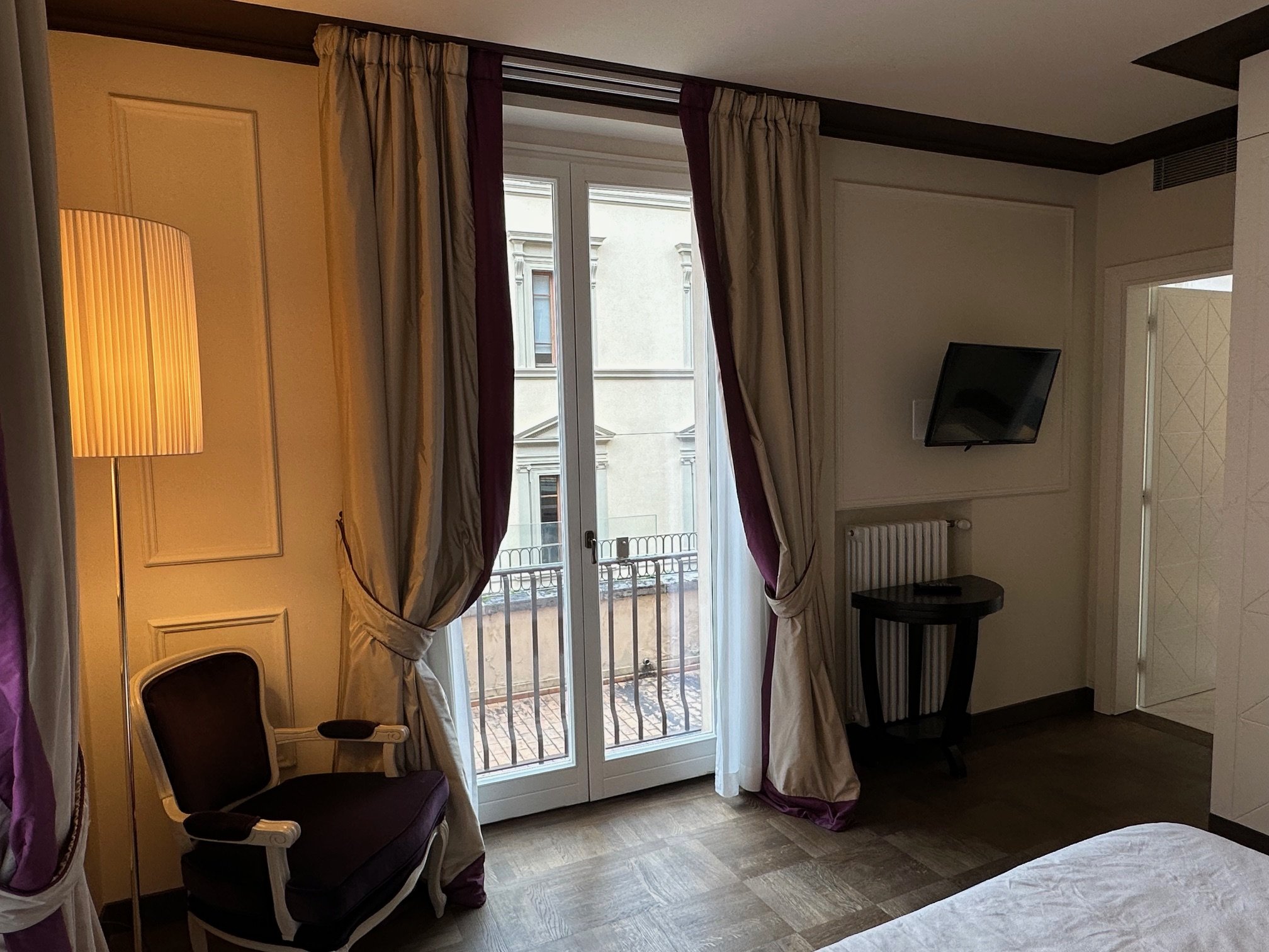 A hotel room with a large window, beige curtains with purple trim, a black side table, a wall-mounted TV, white walls with molding, a floor lamp with a yellow shade, and a chair with dark upholstery.