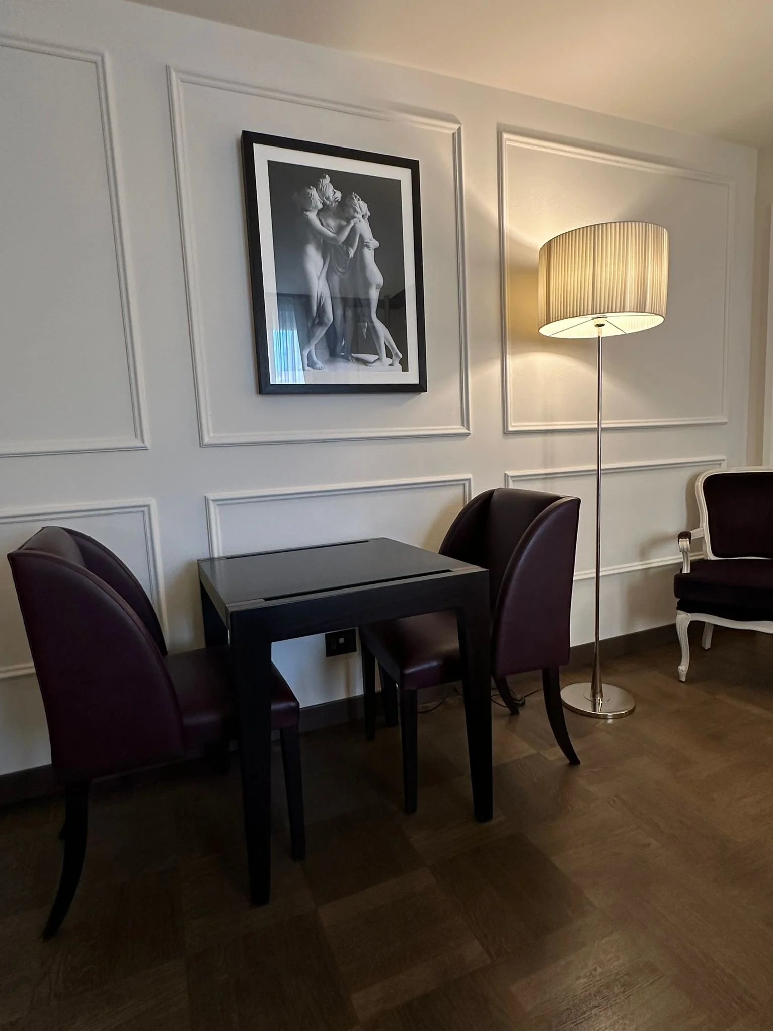 A room with a black table surrounded by purple chairs, a framed black and white artwork of two men, a tall floor lamp with a striped shade, and a white chair against a wall with decorative paneling.