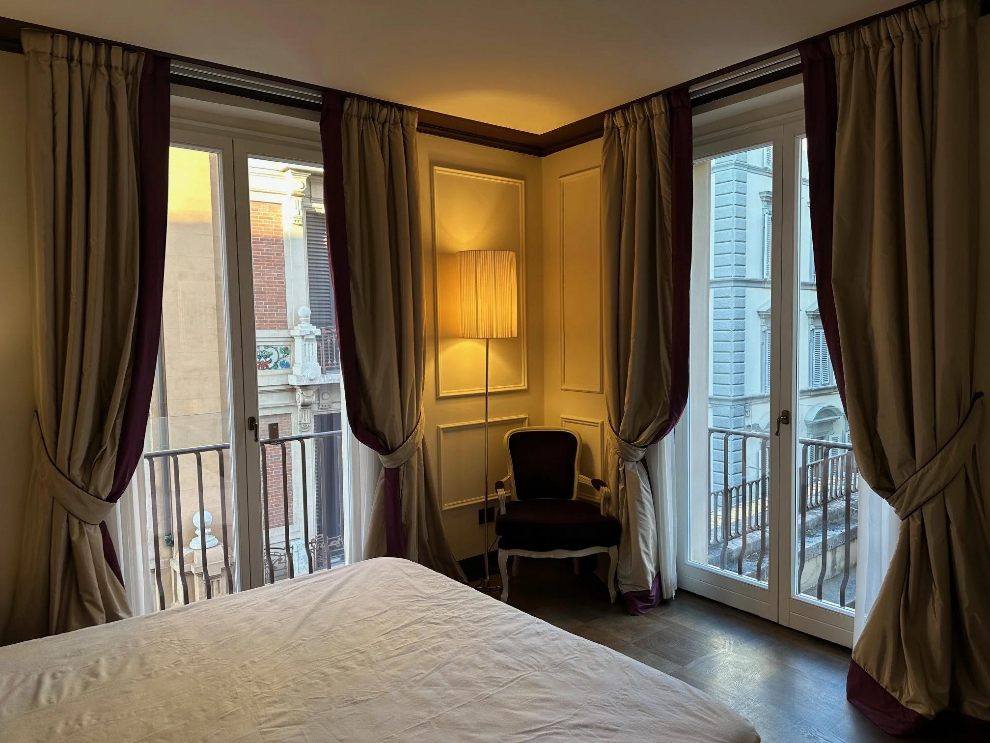 Hotel room with floor-to-ceiling windows, beige curtains with maroon accents, a lamp, a chair, and part of a bed with white bedding.