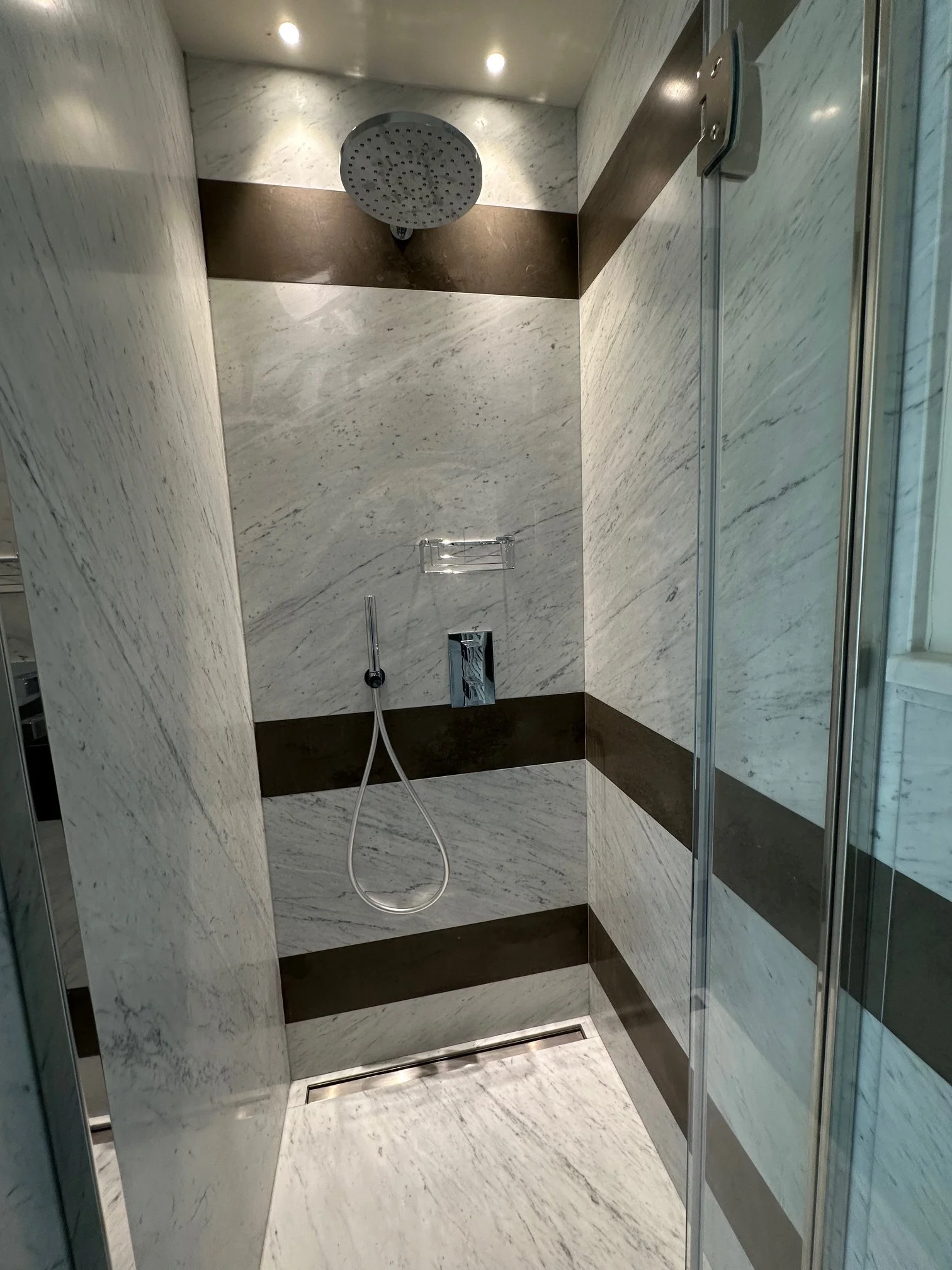 A modern shower with a rain showerhead, marble walls, and a handheld shower. The design features beige and dark brown horizontal bands of marble, with a glass door on the right.