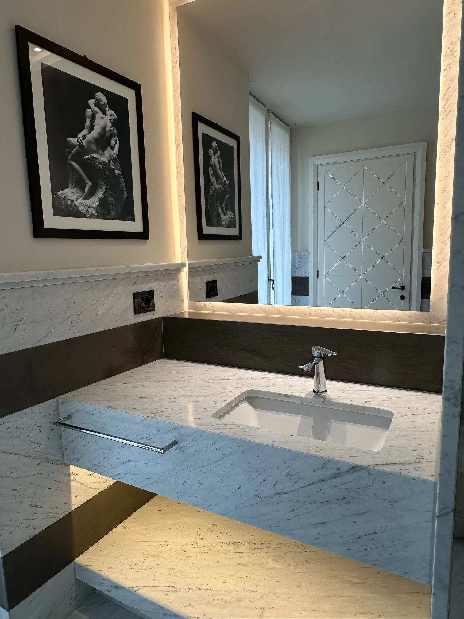Close-up of a bathroom mirror reflecting black-and-white framed photographs of classical sculptures hanging on the wall. The bathroom features marble countertops and a modern faucet.