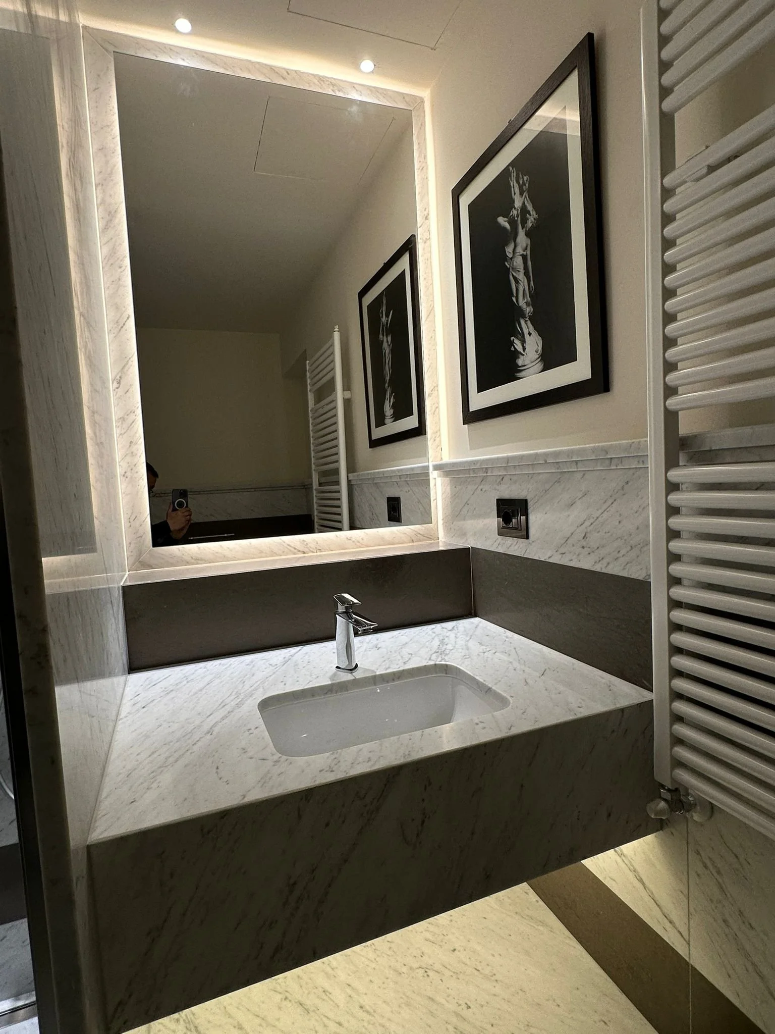 A bathroom sink with a marble countertop and a rectangular mirror above it. To the right, there is a white heated towel rack and black-framed black-and-white artwork on the wall. The reflection shows a person taking the photo and some ceiling lights.