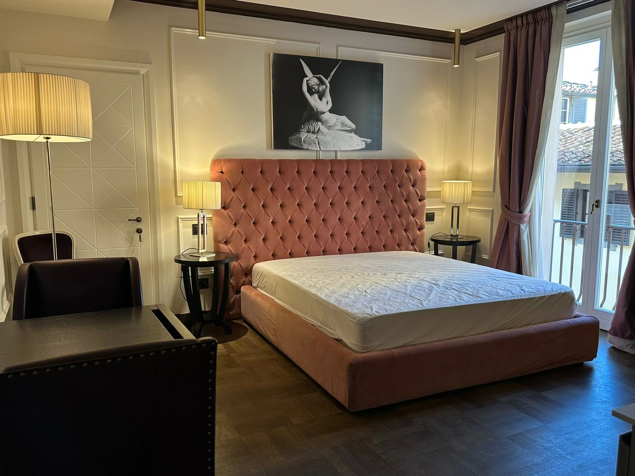 A bedroom with a pink tufted headboard, two bedside tables with lamps, a framed black-and-white artwork of a ballet dancer on the wall, and a large window with curtains showing rooftops outside.