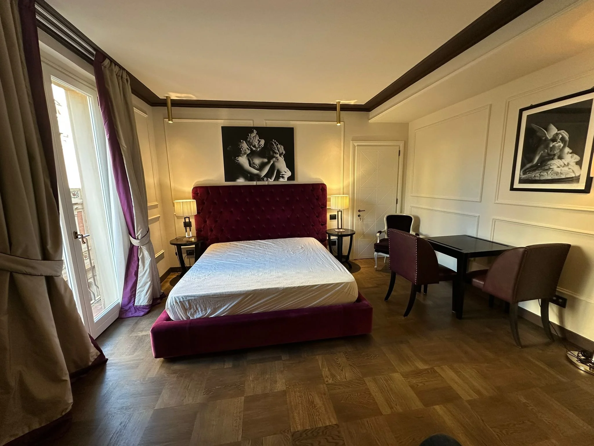 A bedroom with a large bed featuring a red velvet headboard, flanked by two bedside tables with lamps, a black and white artwork of classical sculptures above the bed, a desk with chairs, and windows with cream curtains and purple accents.