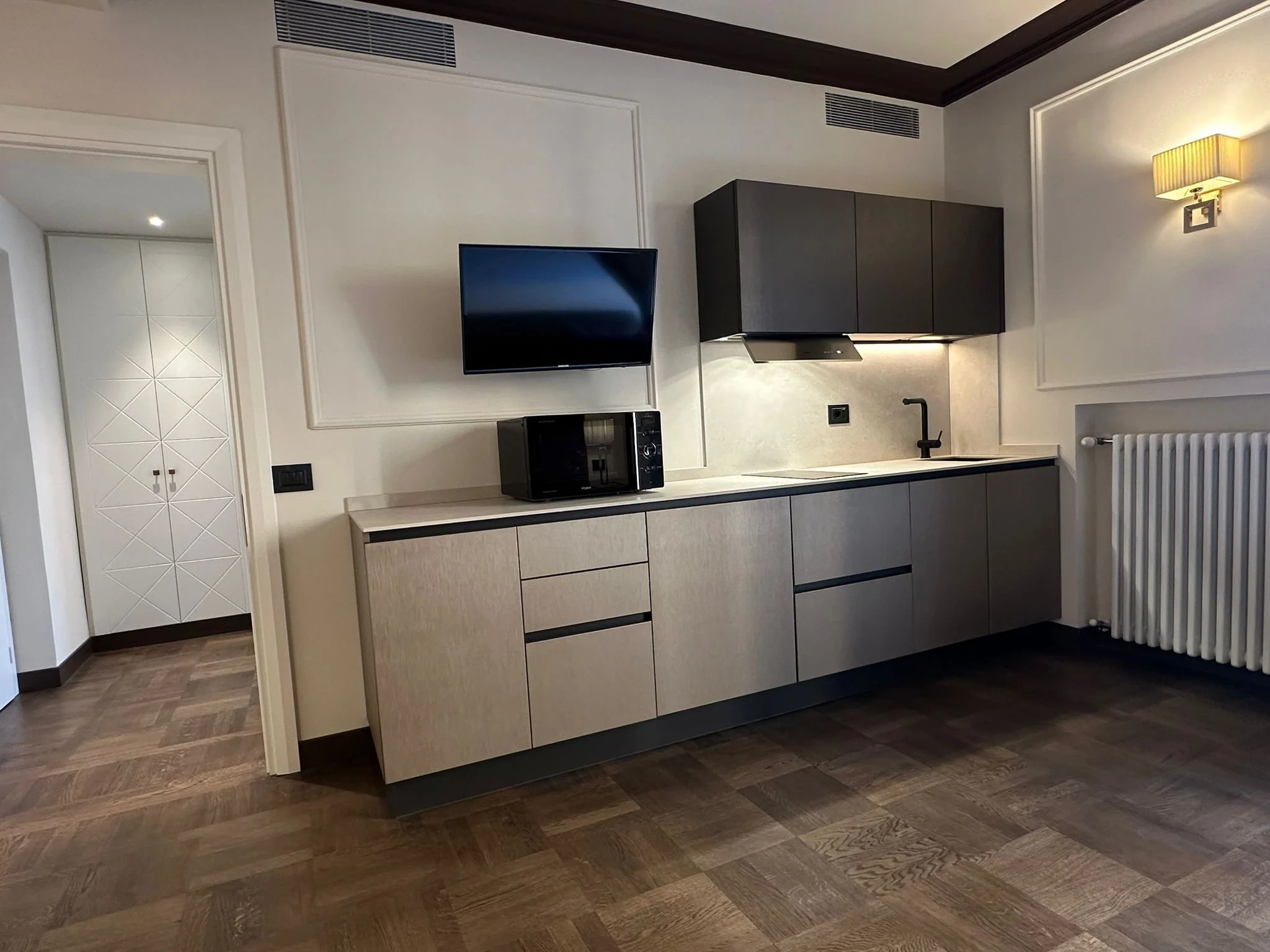 Interior of a modern kitchen with beige cabinets, a black microwave, a black stove hood, a black faucet, and a flat-screen television mounted on the wall. There is a white radiator and dark wood floors.