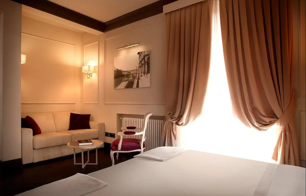 A bedroom with a large window with purple and white curtains, a bed with a mauve velvet headboard, a black and white artwork of three classical statues on the wall, a black side table with a lamp, a white door, and a tall beige floor lamp.