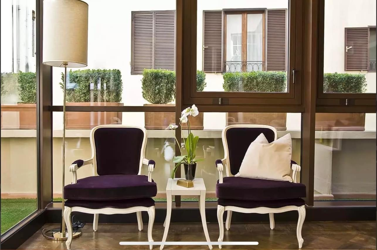 Two black and white armchairs with white pillows and a small white table with a potted orchid between them, in front of a glass wall with a view of a patio with shrubs and window shutters.