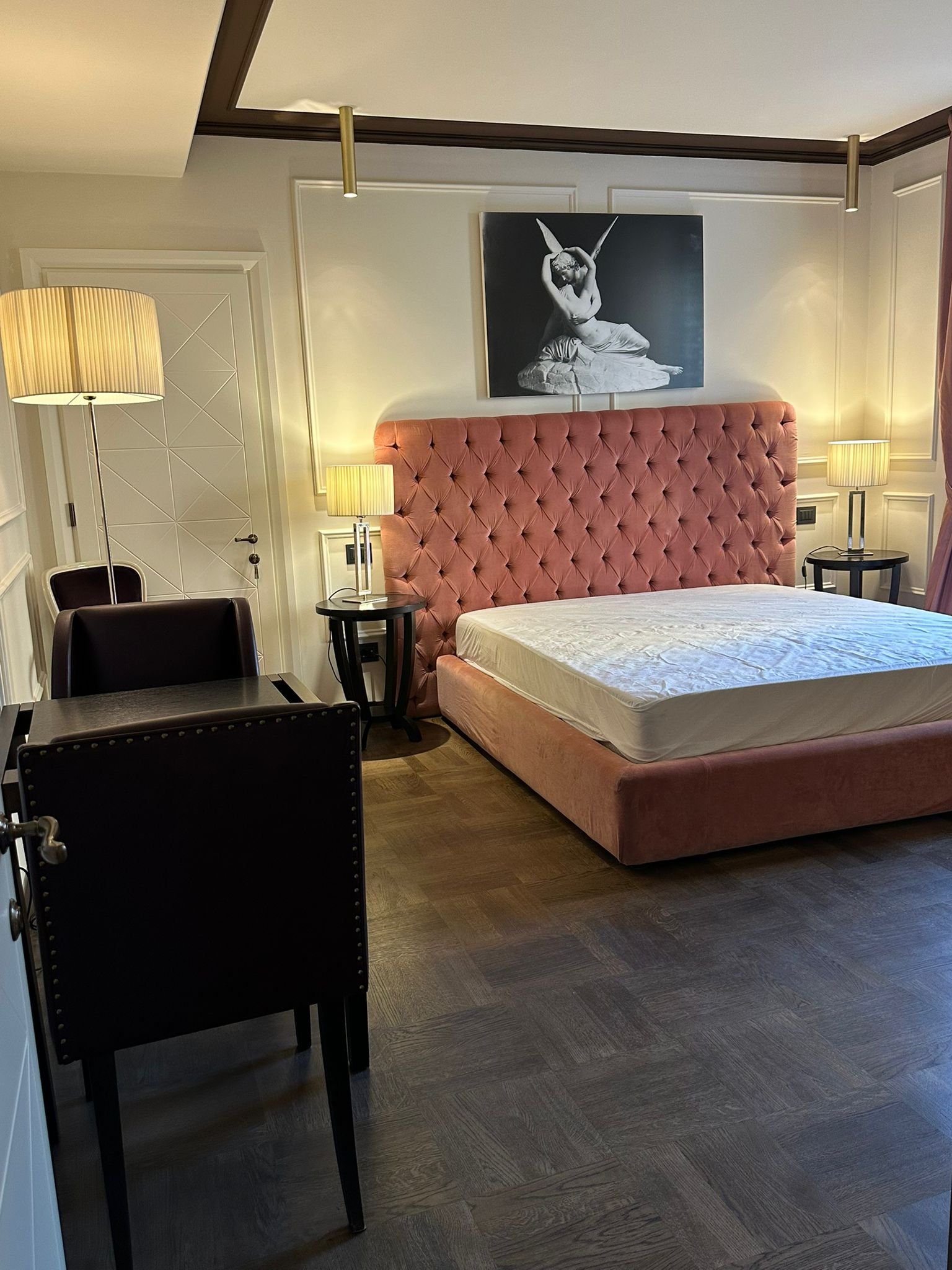 A hotel room with a large bed with a pink, tufted headboard, two nightstands with lamps, a painting of a woman with butterfly wings on the wall, a tall floor lamp, a chair, and a small black table.