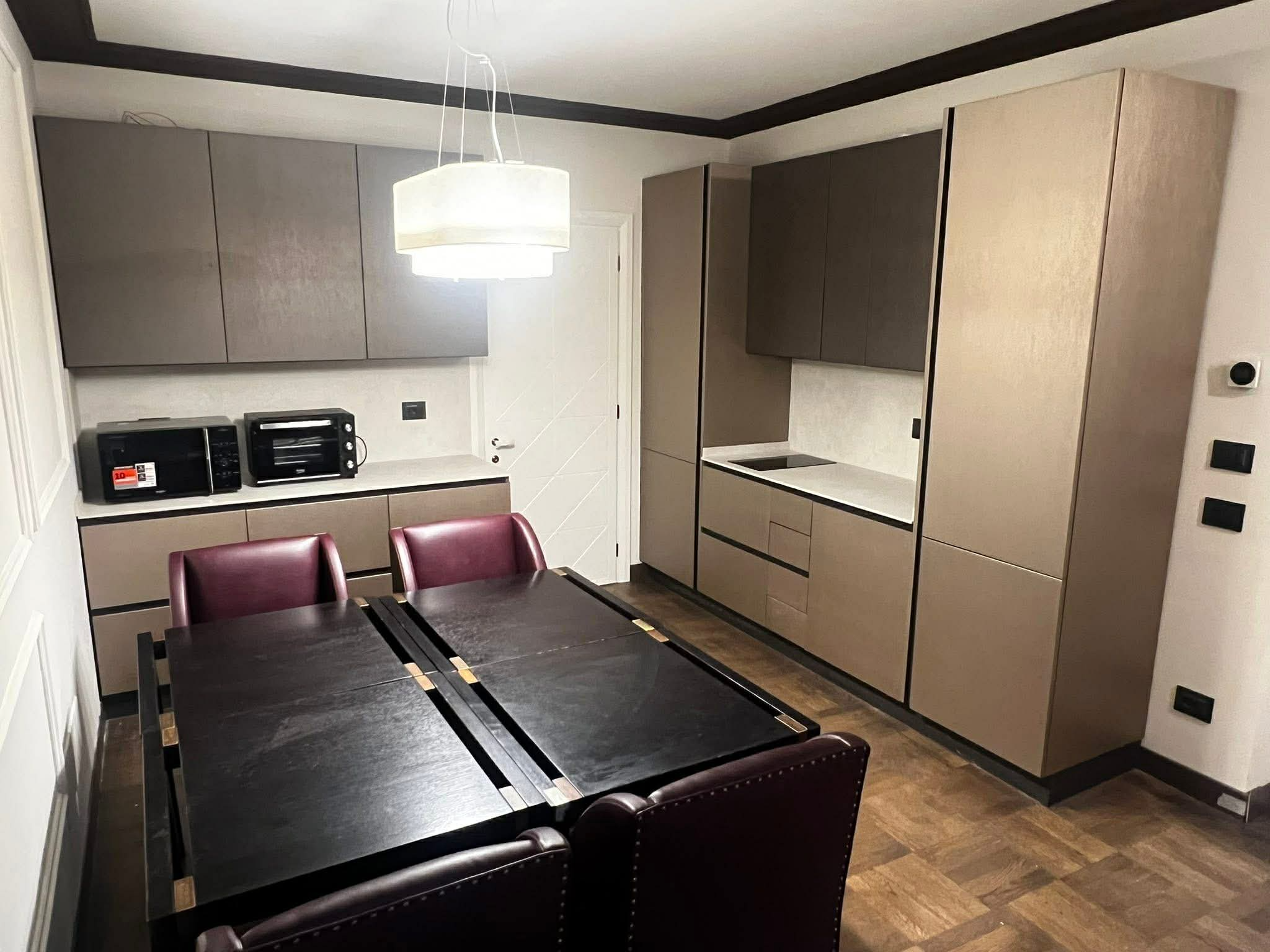 A modern kitchen with beige and gray cabinets, black countertops, a built-in stovetop, a microwave, and a small oven. There is a dining table with four dark red chairs. A hanging white pendant light is above the table.