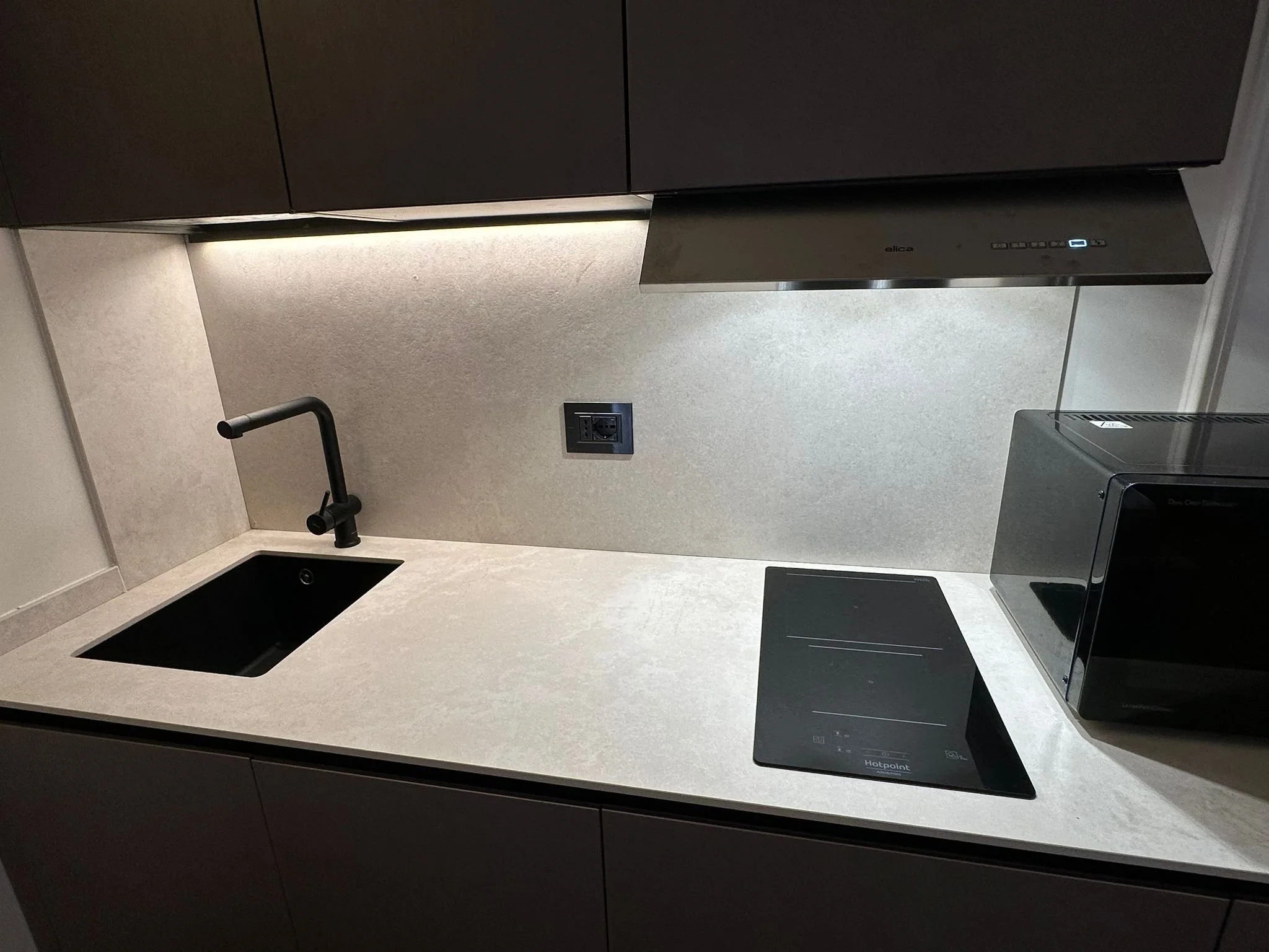 Modern kitchen countertop with black sink, black faucet, beige backsplash, black electrical outlet, stainless steel range hood, black cooktop, and Black hotpoint appliance.
