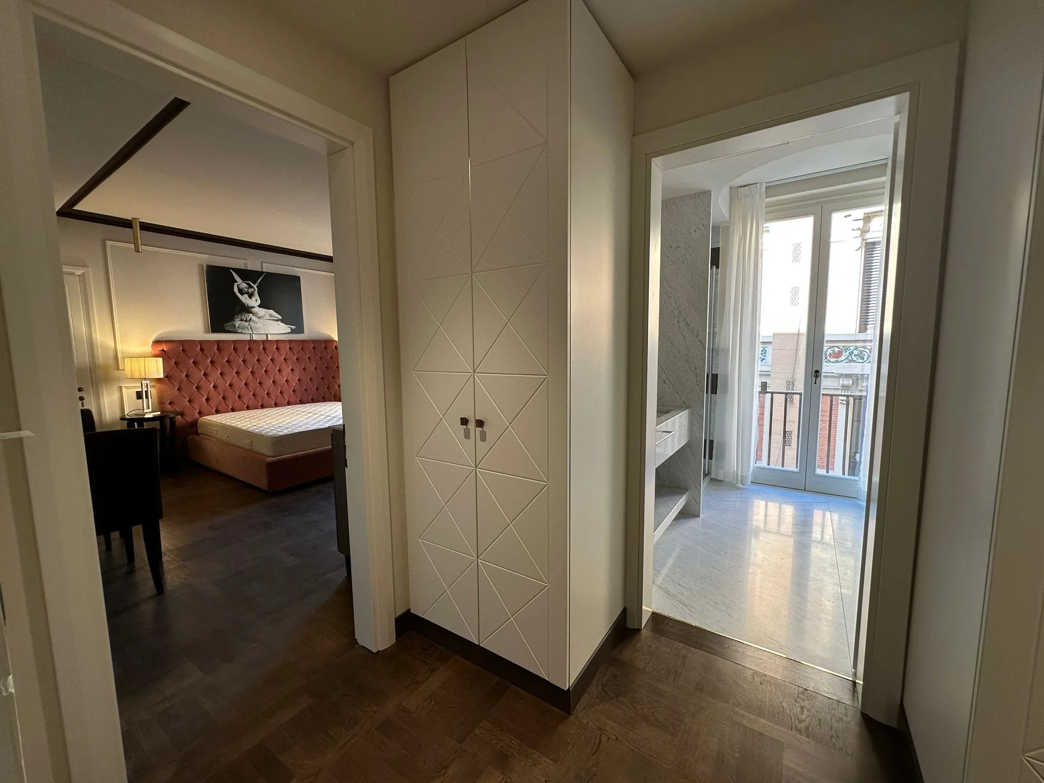 View from the interior of an apartment showing a bedroom with a bed, nightstand, and lamp, and a balcony with a glass door. The hallway has a built-in wardrobe with geometric patterns on its doors.