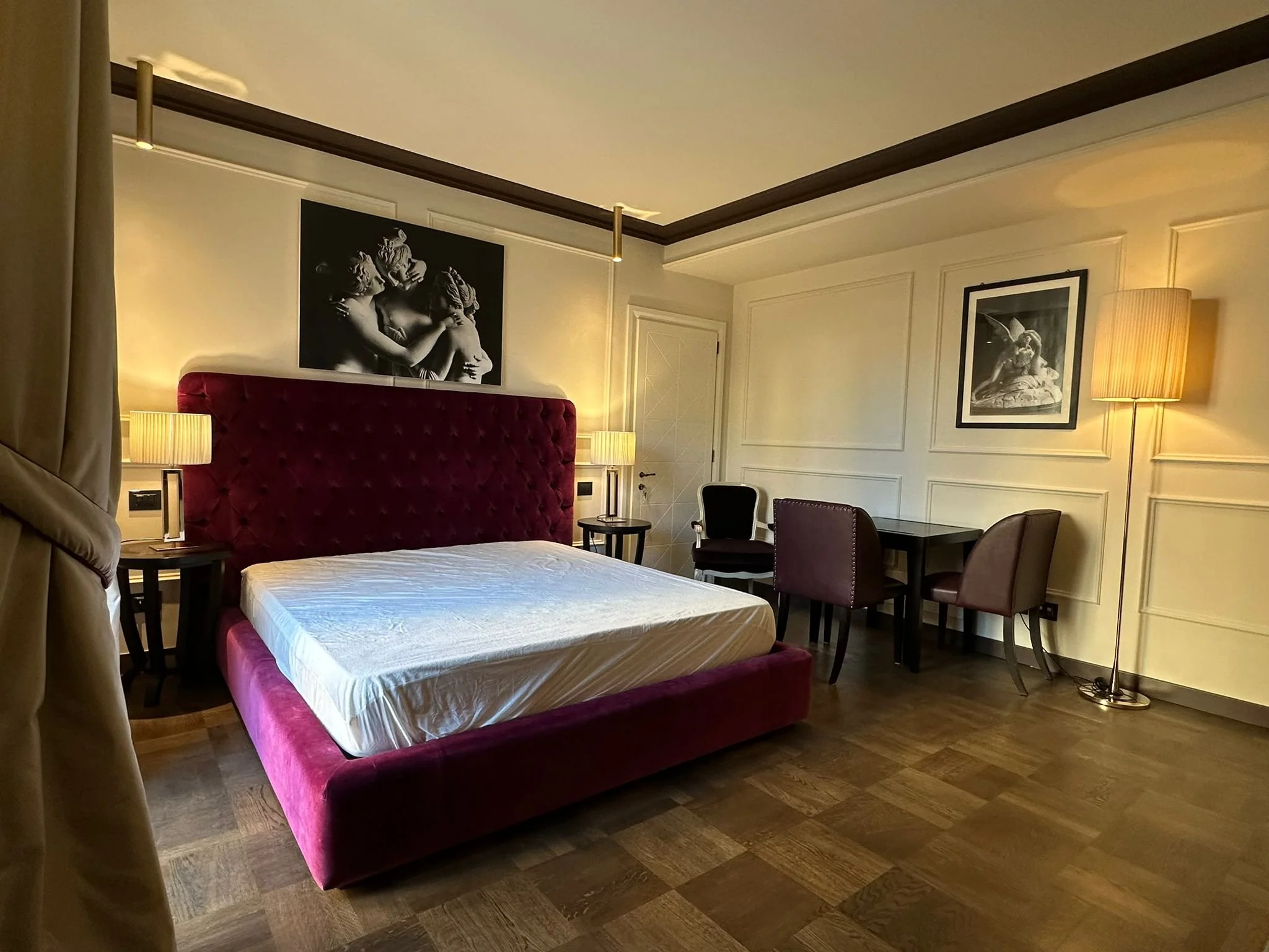 A hotel room with a plush red velvet bed, black and white artwork of cherubs above it, and dark wooden flooring. There are lamps on nightstands, a desk with chairs, and a framed picture of a sculpture on the wall.