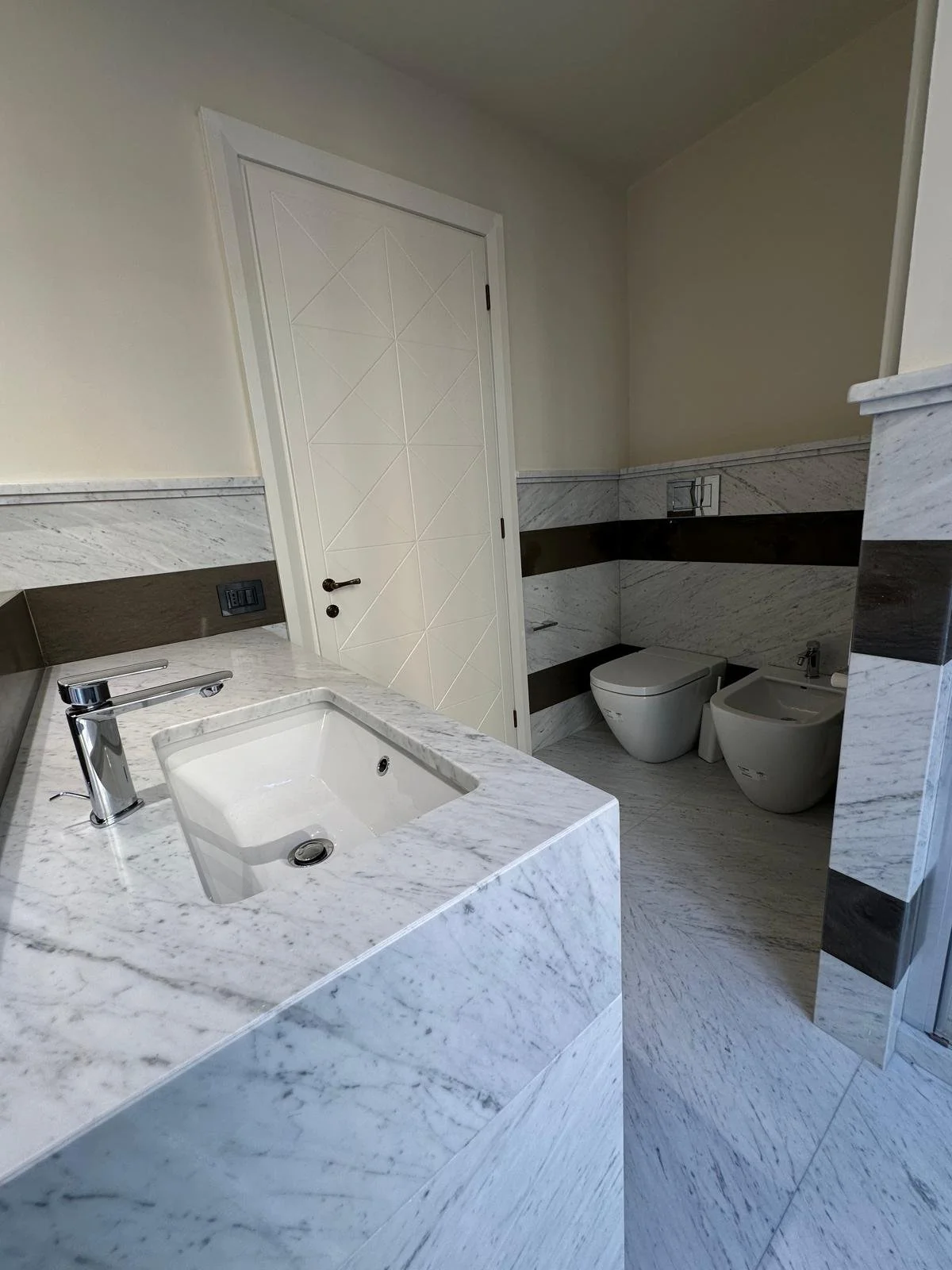 Modern bathroom with marble countertops, a white sink, chrome faucet, toilet, and bidet. The walls have marble and dark stripes, and a white door is visible.