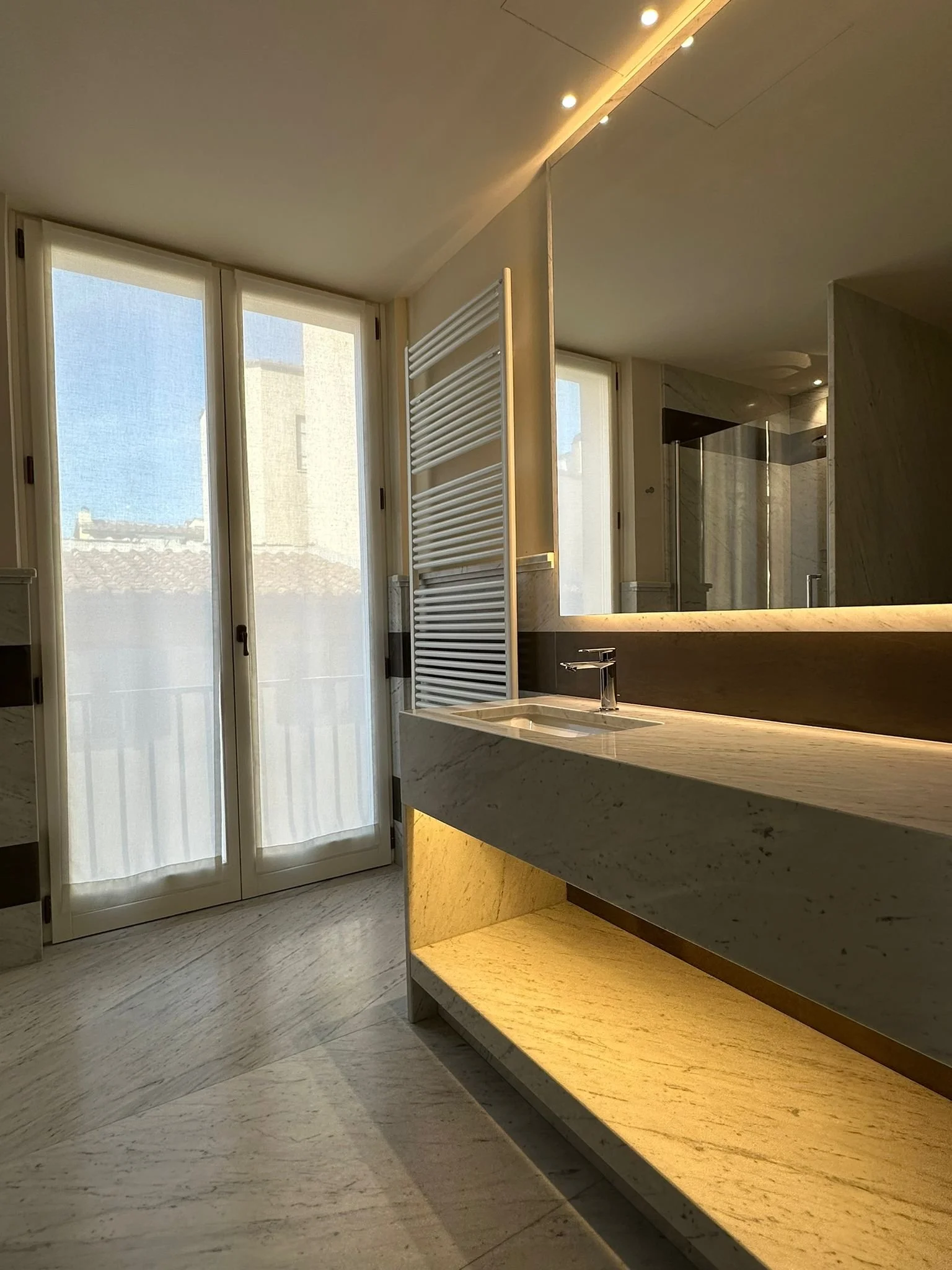 Interior view of a modern bathroom with large windows, a marble countertop with an integrated sink, a tall white radiator towel warmer, and warm, neutral lighting.