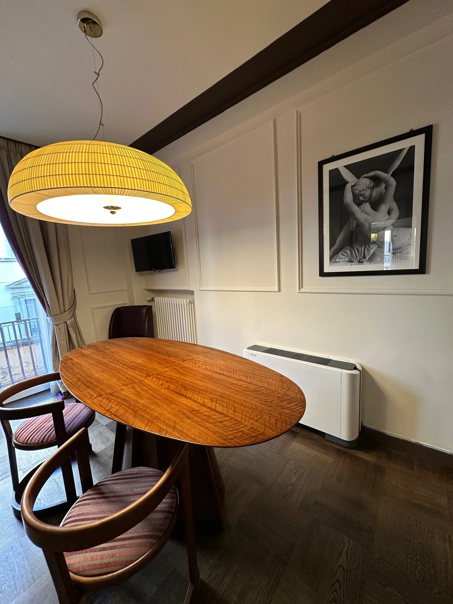 A dining room with a wooden oval table, six chairs with striped cushions, a large yellow pendant light hanging from the ceiling, a wall-mounted television, framed black-and-white artwork of a embrace, and a window with curtains.