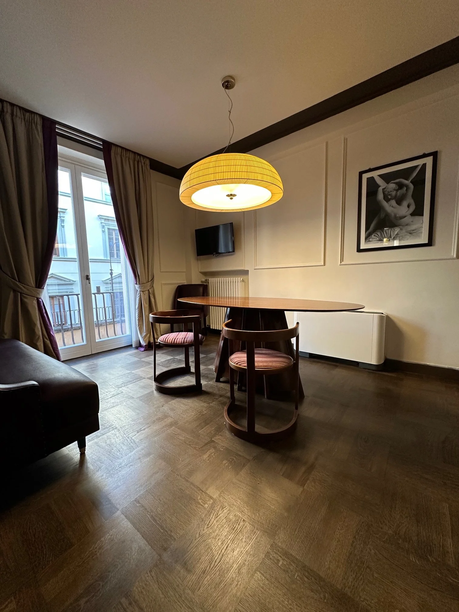 A cozy dining area with three wooden chairs, a round table, and a large yellow hanging pendant light. There are beige curtains on a glass door leading outside, framed black-and-white artwork on the wall, a small flat-screen TV, a white radiator, and 