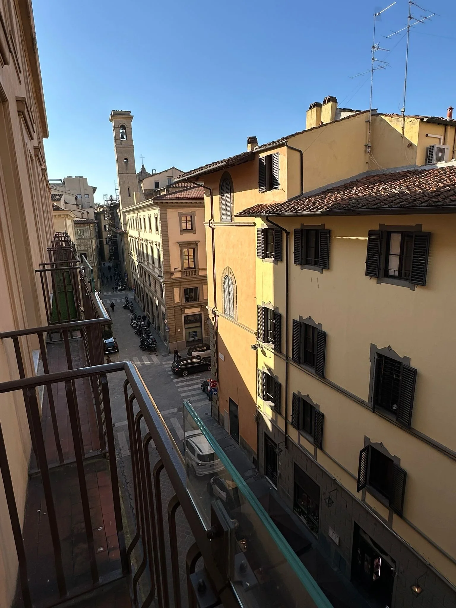 View from a balcony of a narrow city street lined with yellow and beige buildings, with a church bell tower in the distance, under a clear blue sky.