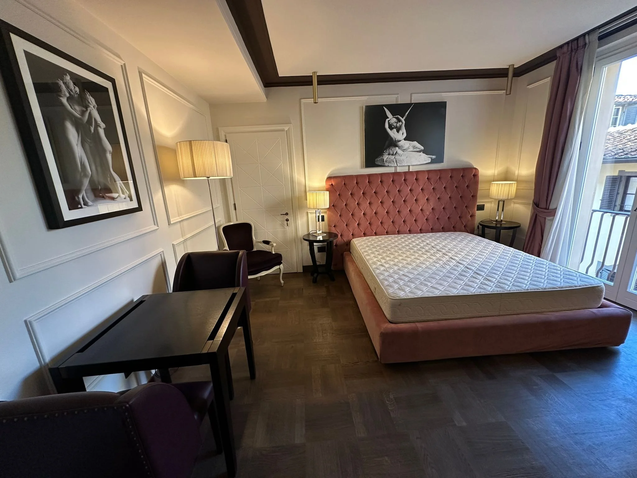 A bedroom with a pink tufted headboard, two bedside tables with lamps, a black chair, a dark wooden desk, framed black-and-white photographs on the walls, a white door, and a window with pink curtains showing rooftops outside.