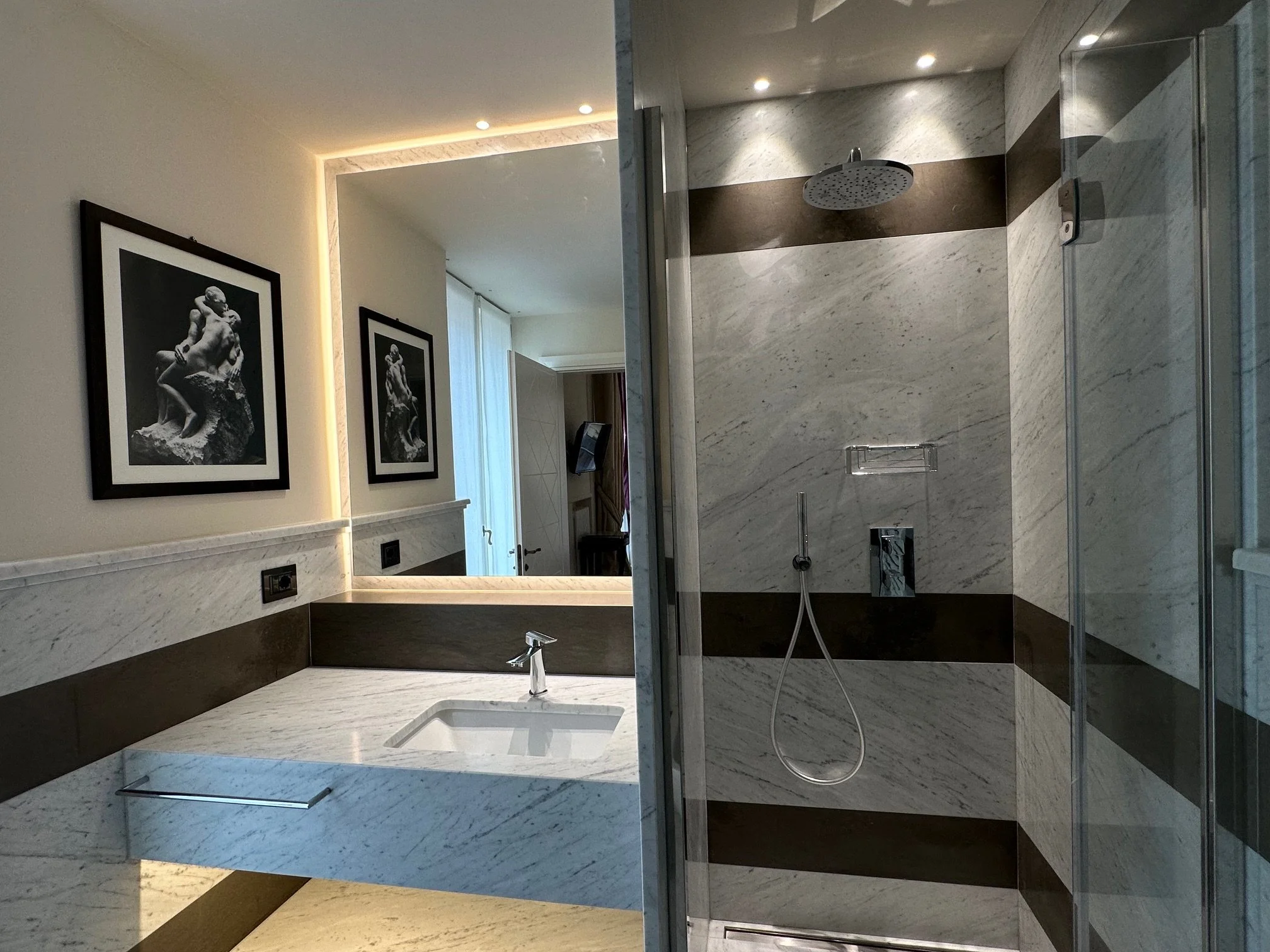 Modern bathroom with marble walls and a walk-in shower, featuring a rainfall showerhead and two framed black and white sculptures on the wall.