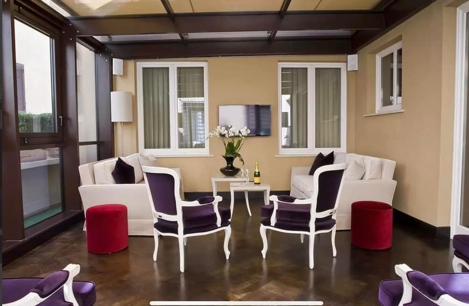 Indoor patio with beige walls, white windows, and dark wood floor. There are two white sofas with black cushions, two black and white armchairs, and two red ottomans. A TV is mounted on the wall, with a large vase of white flowers and a bottle on a s