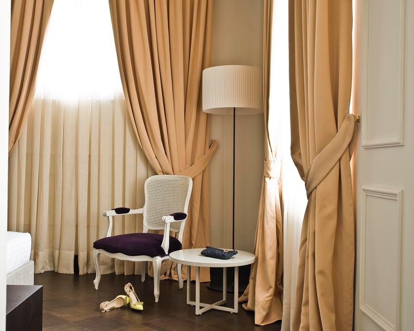 Elegant room with beige curtains, a white armchair with dark purple cushion, a black floor lamp, a small white table with a remote control, a pair of beige high heels, and sunlight coming through the window.