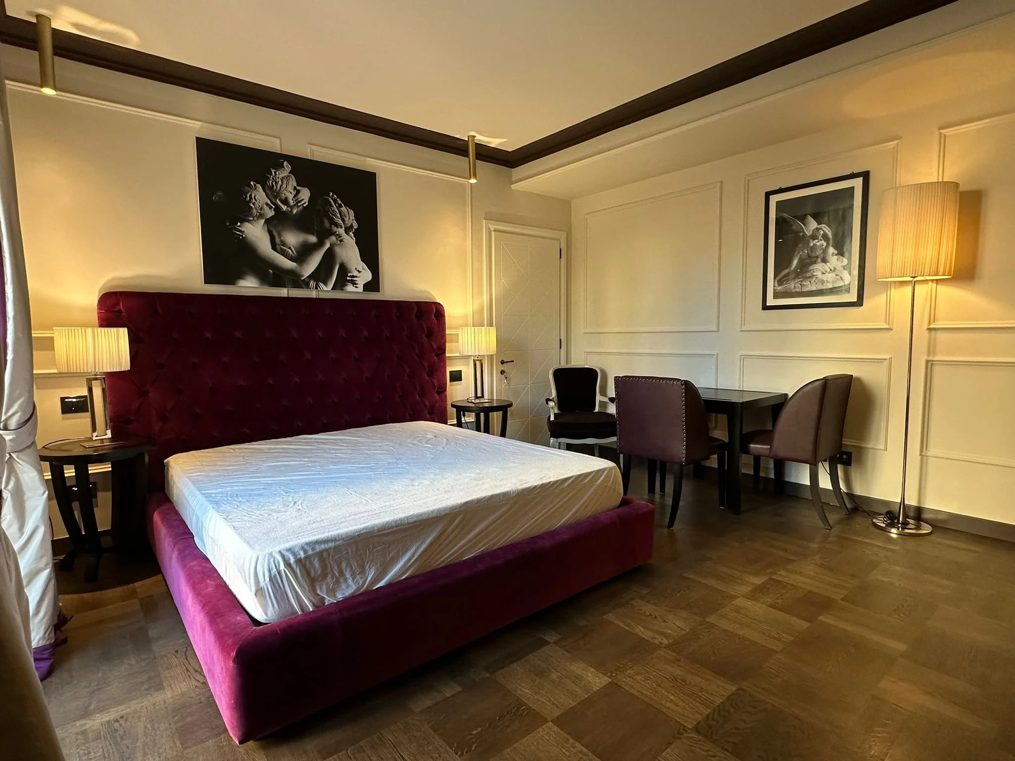 A hotel room with a large bed that has a maroon velvet headboard, side tables with lamps, and a sitting area with chairs and a table. There are framed black-and-white art pieces on the cream-colored walls, and a floor lamp next to one of the chairs.