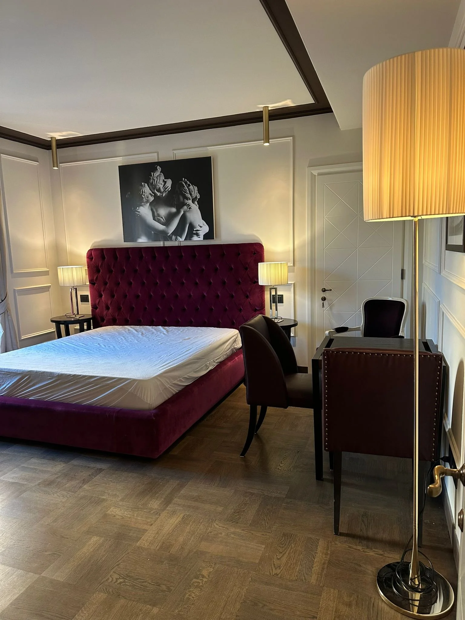 A bedroom with a bed that has a red, tufted headboard, two bedside tables with lamps, a black chair, a large floor lamp, a black and white artwork of classical sculpture on the wall, and wooden flooring.