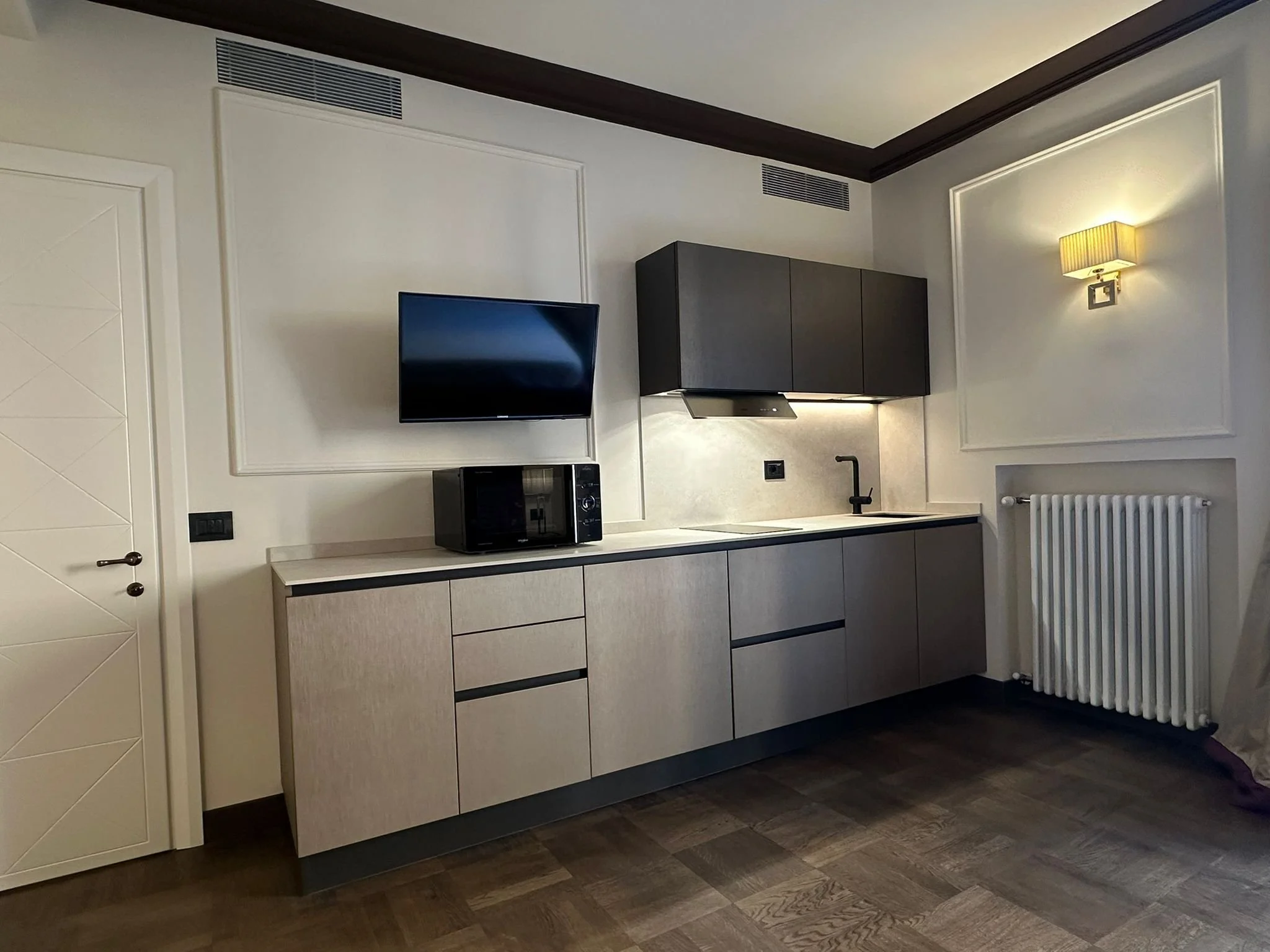 Modern kitchen with beige and black cabinetry, a wall-mounted TV, a microwave, a small countertop, black sink, and a yellow wall lamp.