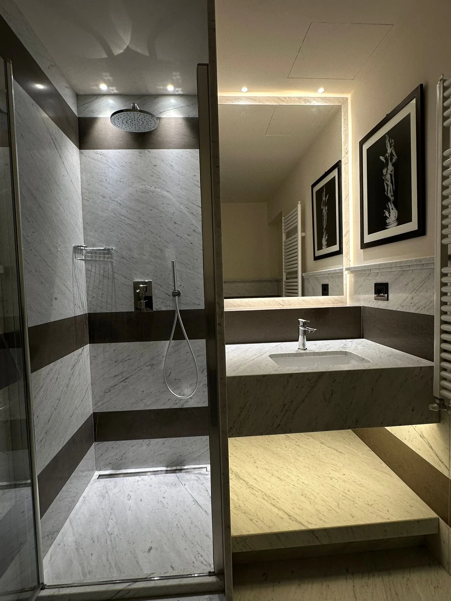 Interior view of a modern bathroom, featuring a shower area with a round rain shower head, marble walls and floors, and two framed black and white photographs of a statue on the wall above a large mirror with backlit lighting.