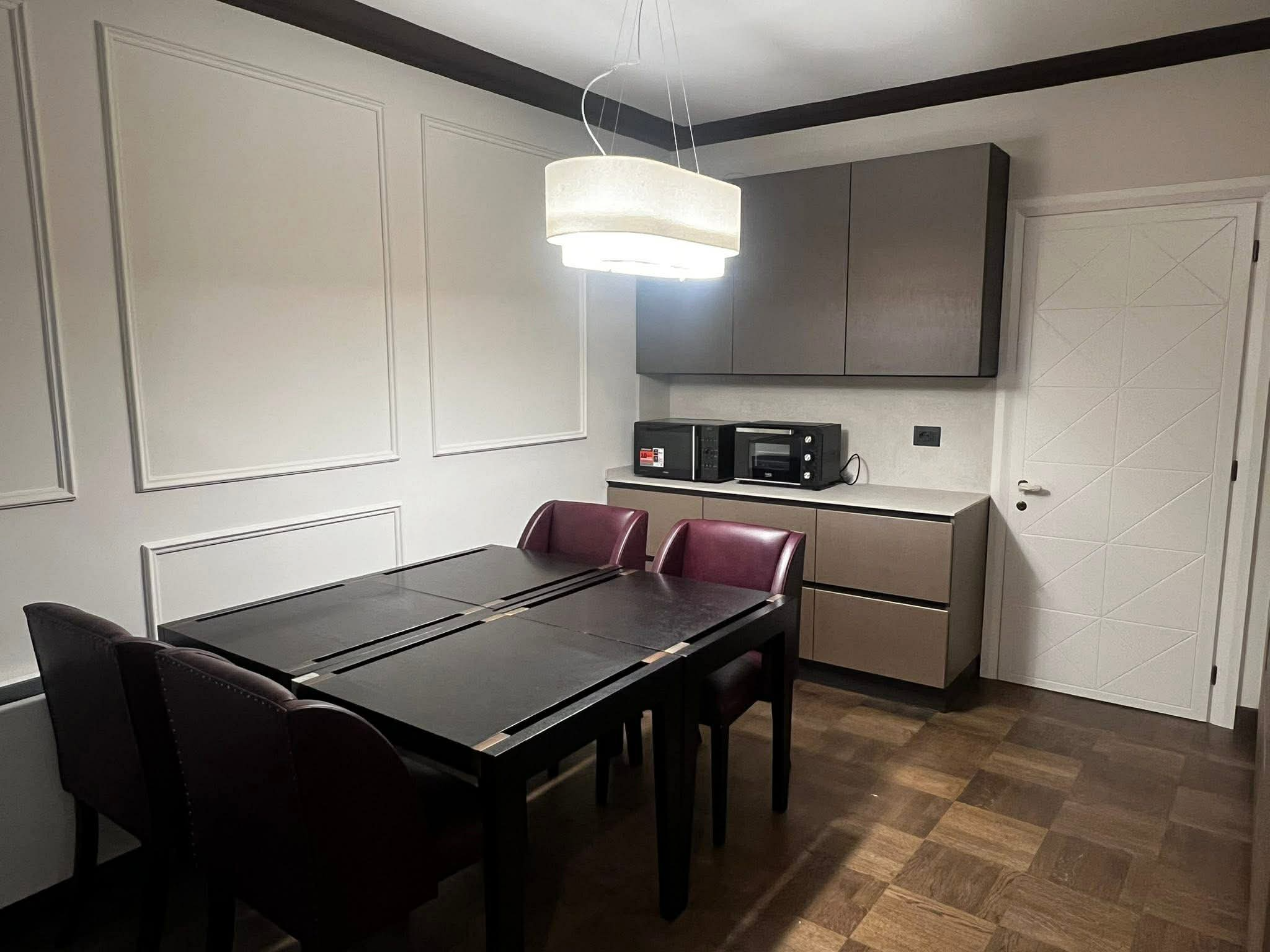 Interior of a modern dining room with a rectangular black table and six burgundy chairs. Cream-colored walls with decorative molding, dark wood trim on the ceiling, and a white door with a diamond pattern. Kitchen appliances including a microwave and