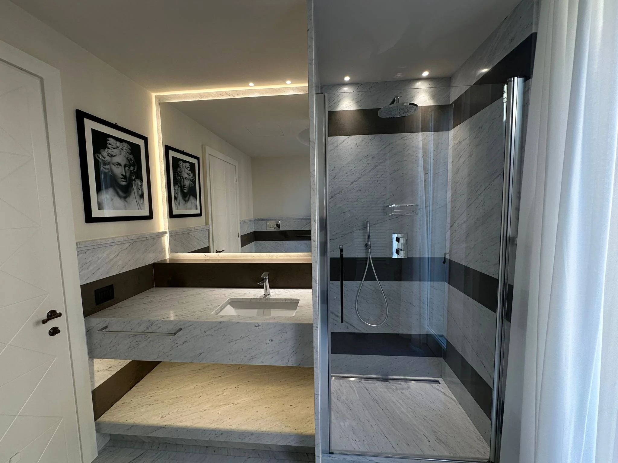 Modern bathroom with marble walls and floor, featuring a walk-in shower with glass doors, a rain showerhead, and black accent stripe. Two black and white framed portraits of a woman adorn the wall.