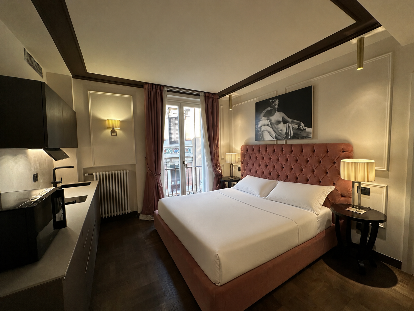 Hotel room with a large bed with a pink tufted headboard, framed artwork above, bedside lamps, a window with curtains, a small balcony, a kitchenette with black cabinets, a sink, and a radiator below the window.