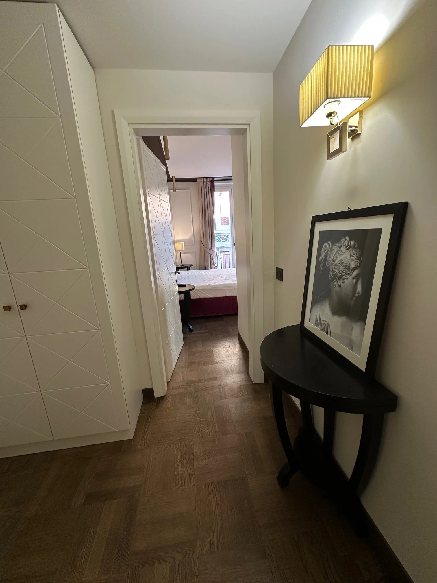 The image shows a hallway with wooden flooring, white walls, a decorative white cabinet with a textured geometric pattern on the left, a black round side table with a framed artwork leaning against the wall, a wall-mounted lamp with a beige pleated s