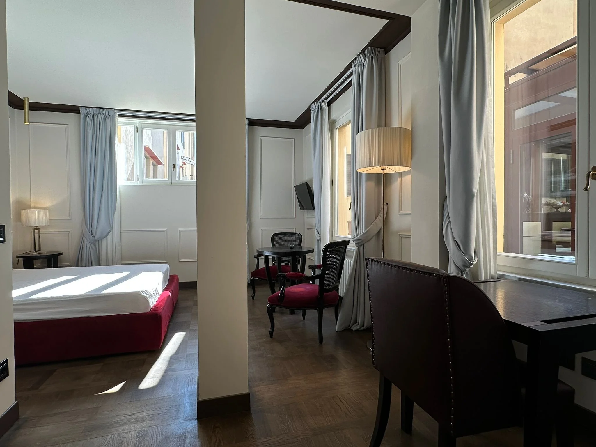 A bright room with multiple windows, white curtains, and wooden flooring. There is a bed with white linens on the left, a small black table with a lamp, a round dining table with four chairs, and various lamps providing warm lighting. The room has a 