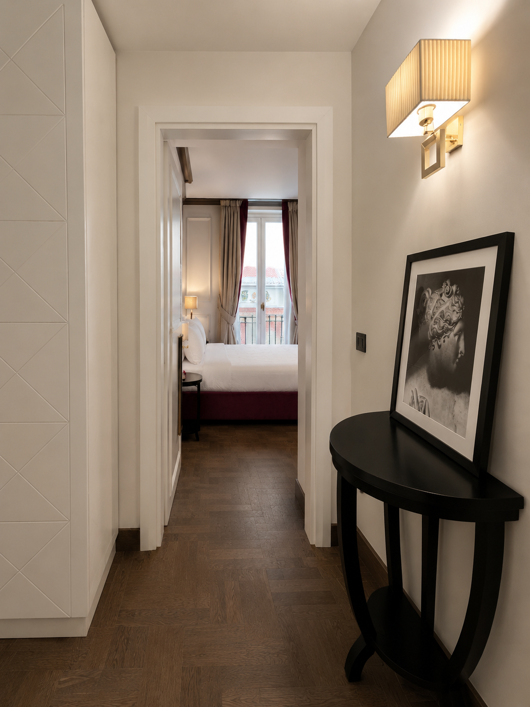 View from a hallway into a bedroom with a window, curtains, bed, and nightstand. There is a framed black-and-white photo on a black table, and a wall-mounted light fixture.