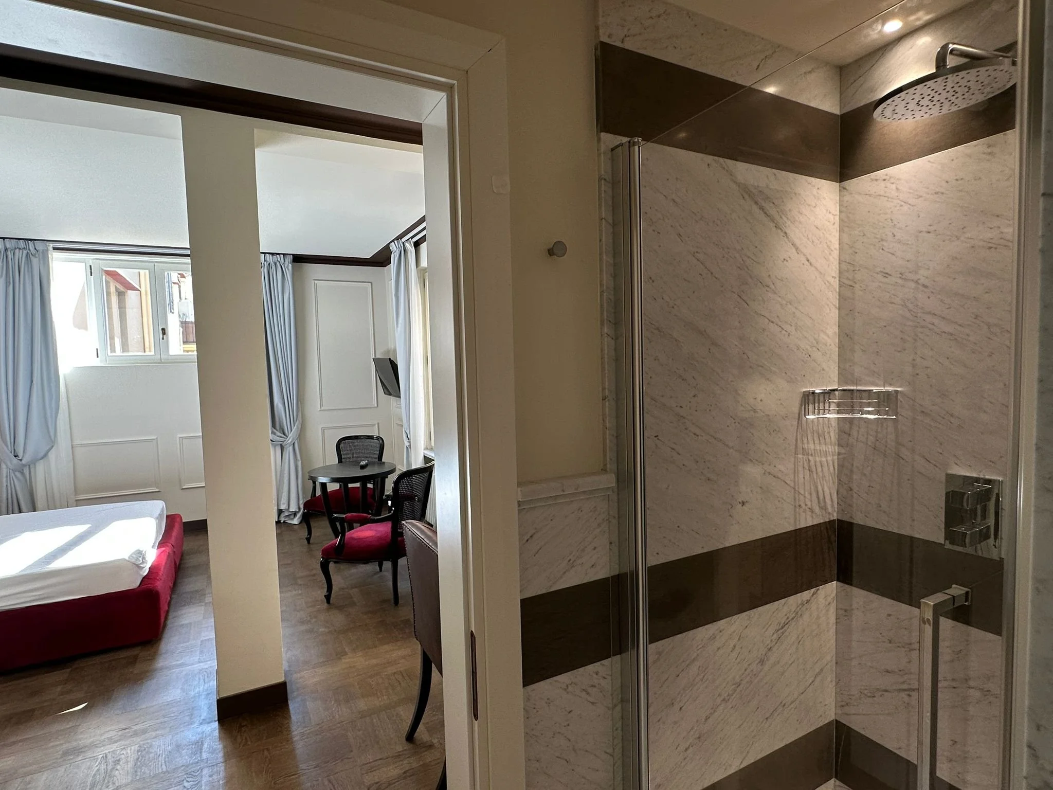 The image shows a hotel room with a sleeping bed, chairs, windows with curtains, and an open shower area with marble walls.