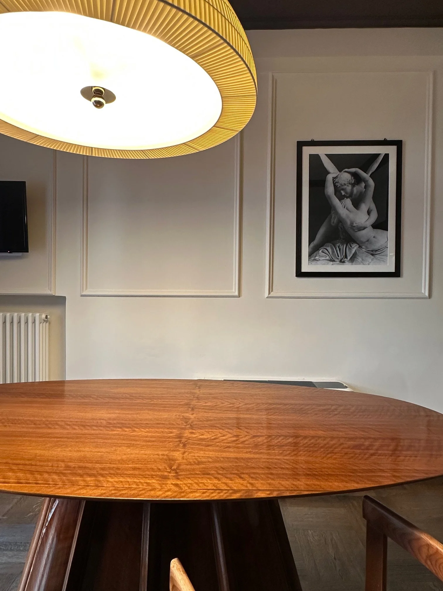 A wooden dining table with a smooth polished surface, surrounded by wooden chairs, is positioned in front of a white paneled wall. A large, modern ceiling light fixture with a circular design and a yellow rim hangs above.