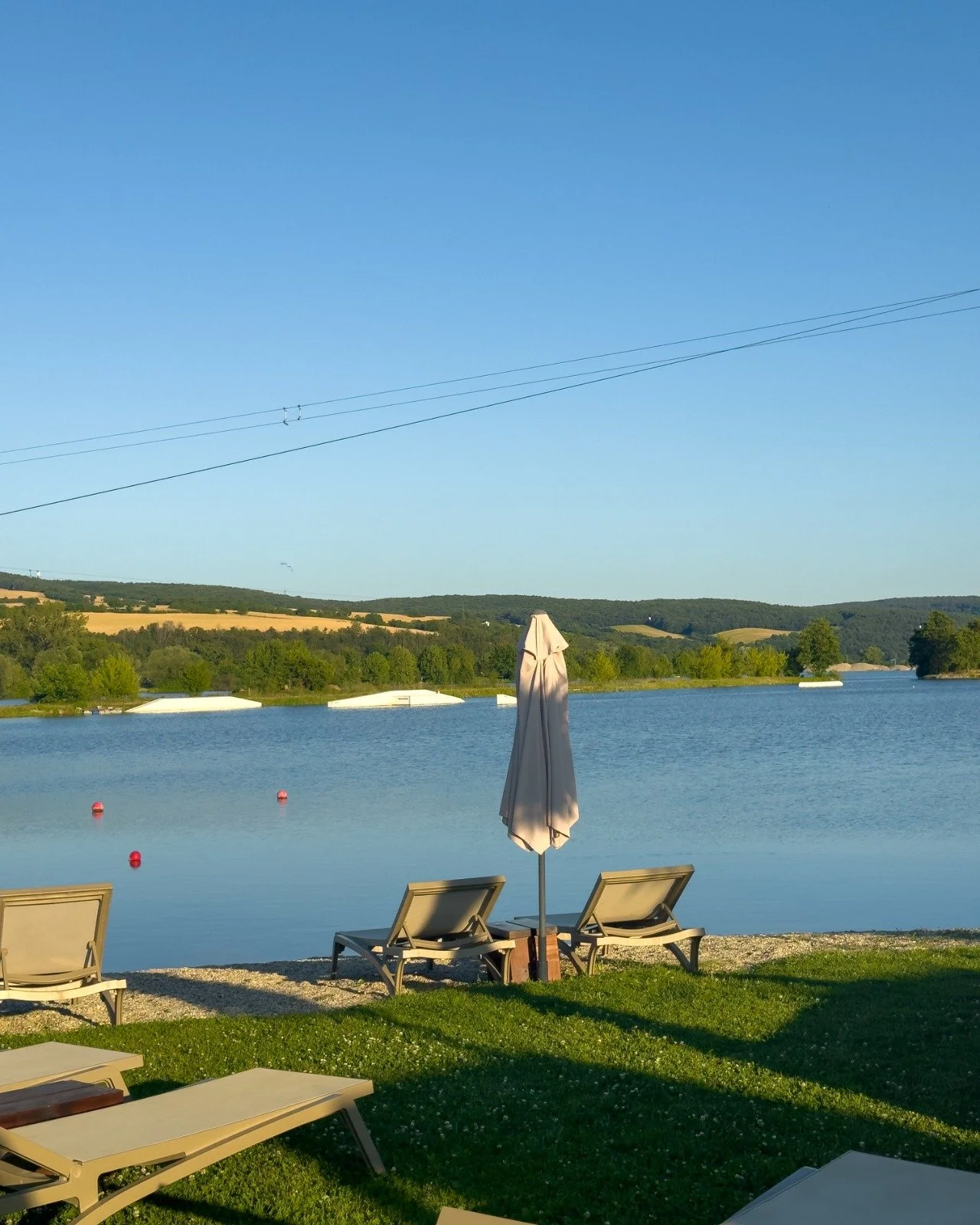 Lakeside scene with a river or lake, lounge chairs on a grassy area, a closed umbrella, trees across the water, hills in the background, and a clear blue sky.