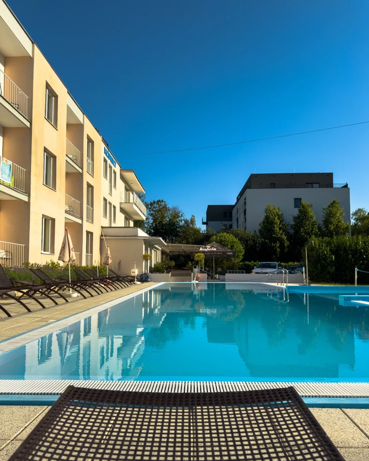 Swimming pool with lounge chairs and umbrellas in front of modern apartment buildings under a clear blue sky.