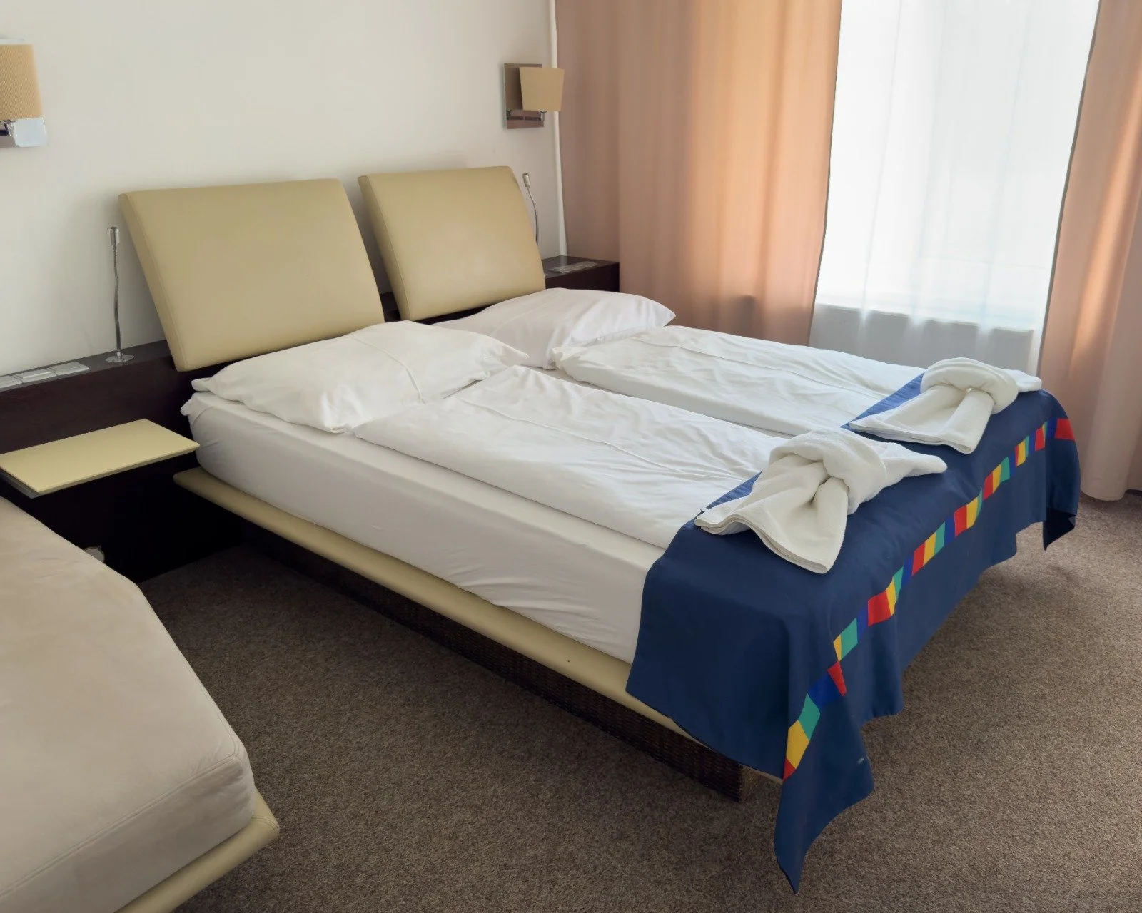 Hotel room with two neatly made beds, folded towels on the bed, bedside lamps, and curtains covering the window.