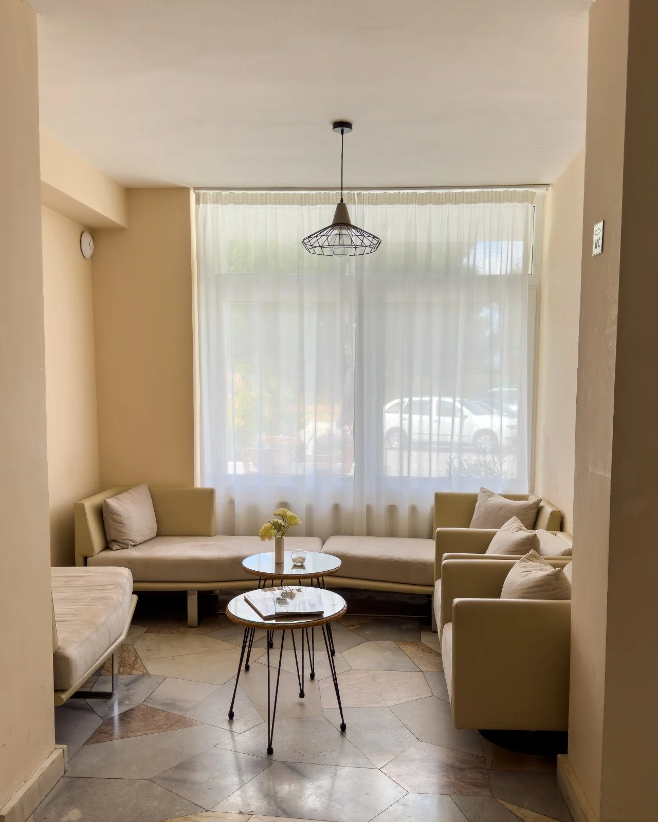 Cozy living room with beige sofas, a white round coffee table with flowers, large window with sheer curtains, and a modern pendant light.
