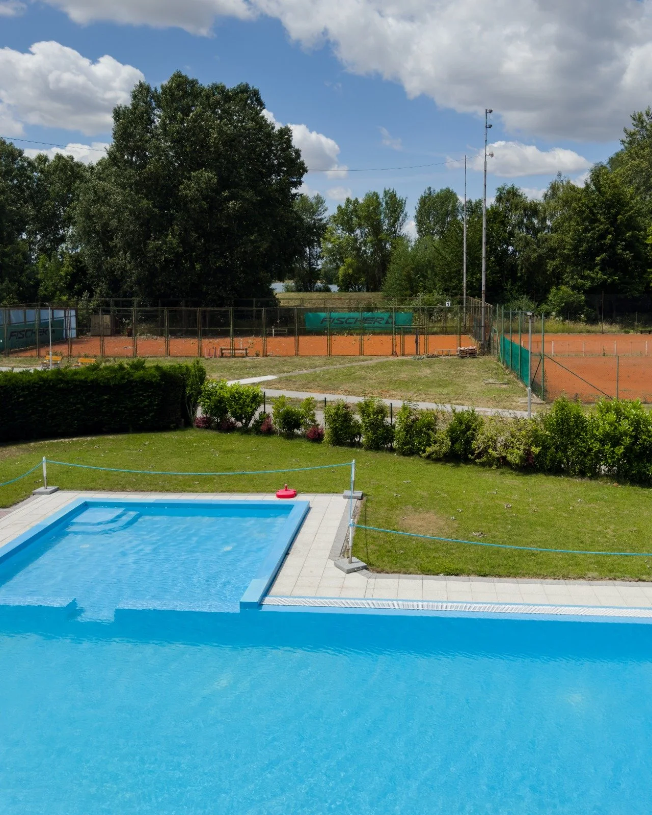 Photo of a swimming pool with a pink pool float, surrounded by a well-maintained lawn and a garden, with tennis courts and trees in the background under a partly cloudy sky.