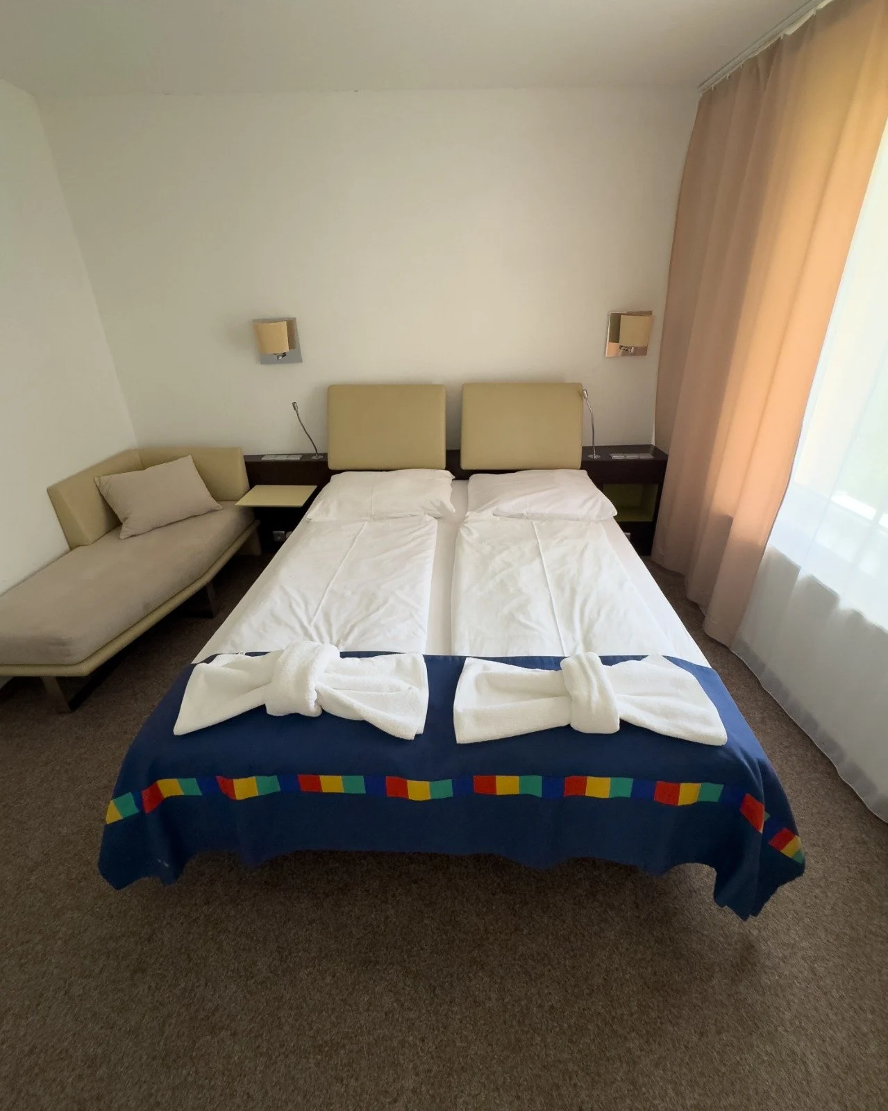 A hotel room with twin beds pushed together, two bath towels tied into bows on the beds, a small couch on the left, a window with curtains on the right, and two wall-mounted lamps behind the beds.