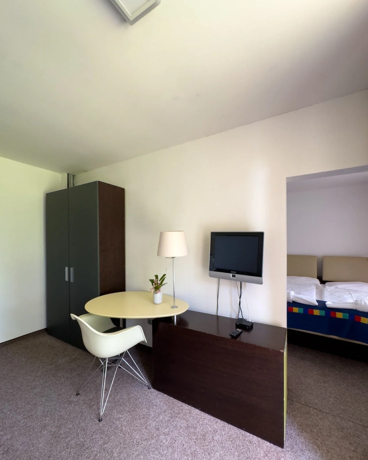Hotel room with a small round table, a white chair, a TV mounted on the wall, a bed with white linens, a dark wardrobe, and a lamp on the table, with a window not visible in the image.