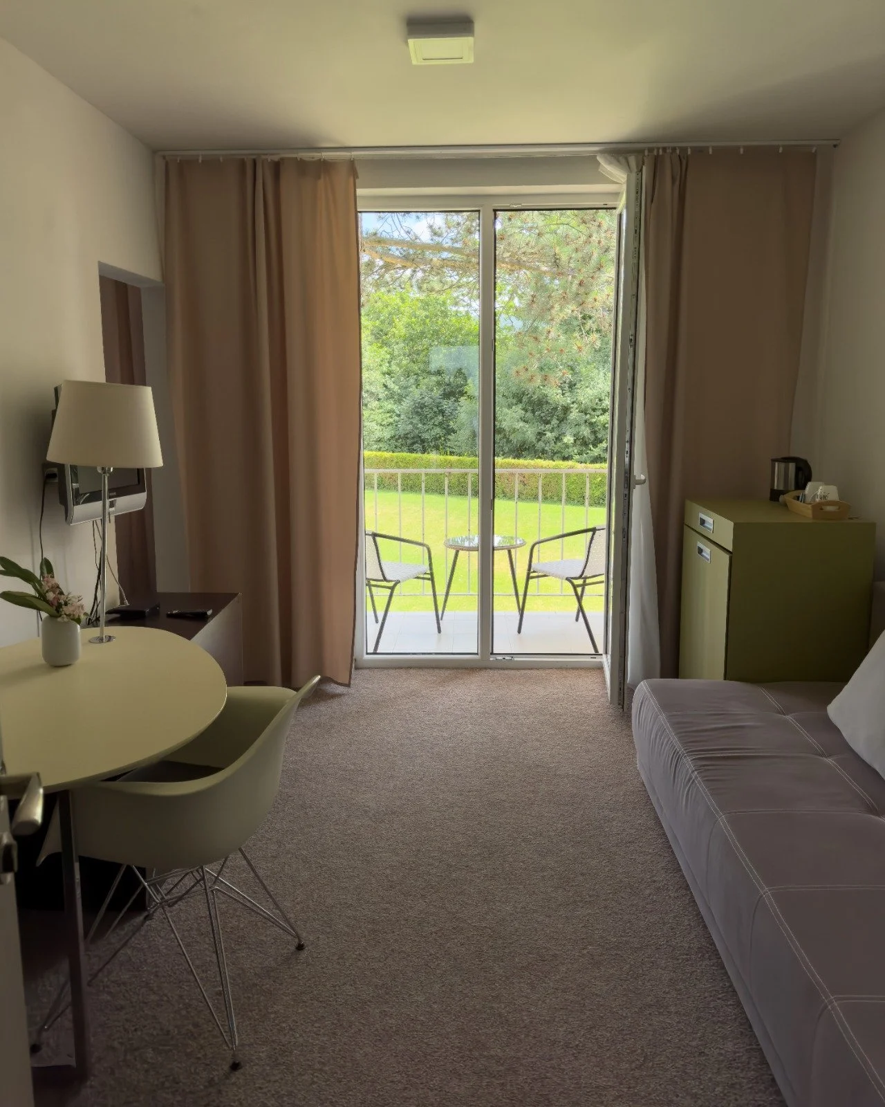 Hotel room with sliding glass door leading to balcony with two chairs and a table, beige curtains, a small green cabinet, a sofa, a round dining table with chairs, and a view of trees and a lawn outside.