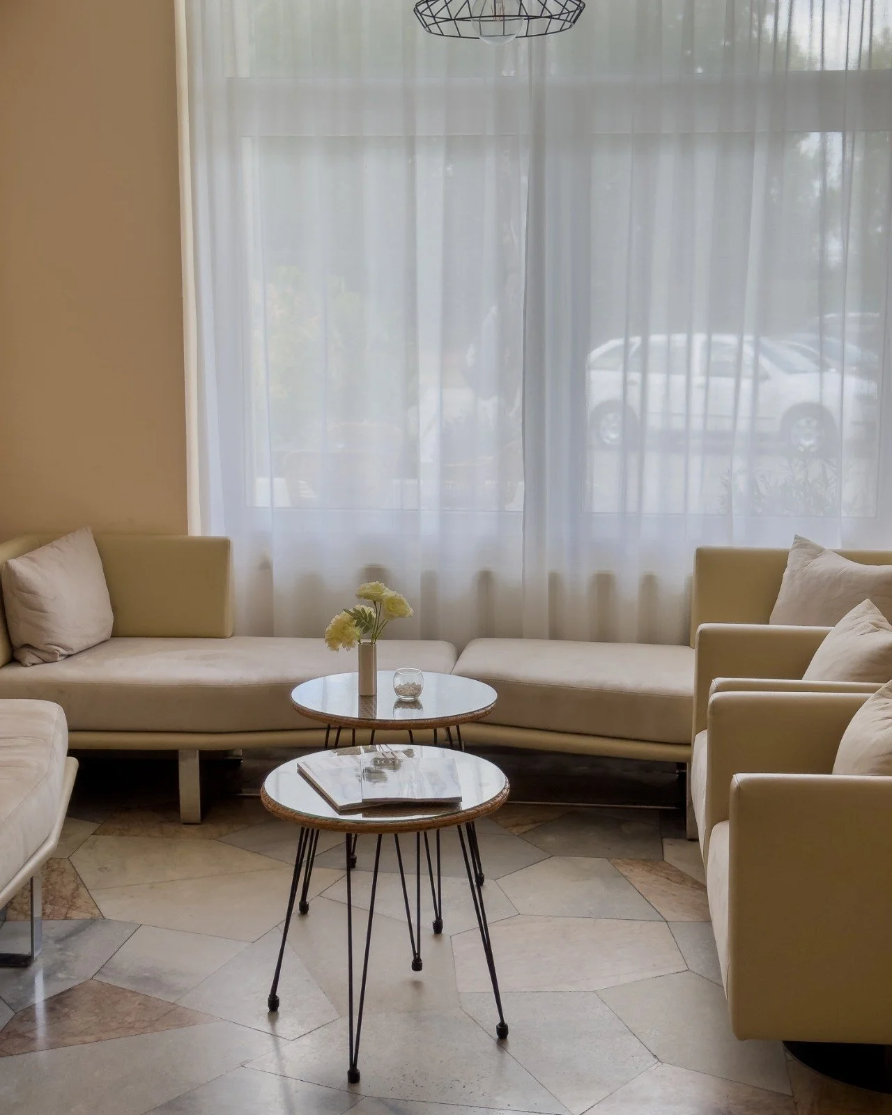 Living room with a large window with sheer curtains, cream-colored sofas, two round coffee tables, a vase with flowers, and a chandelier